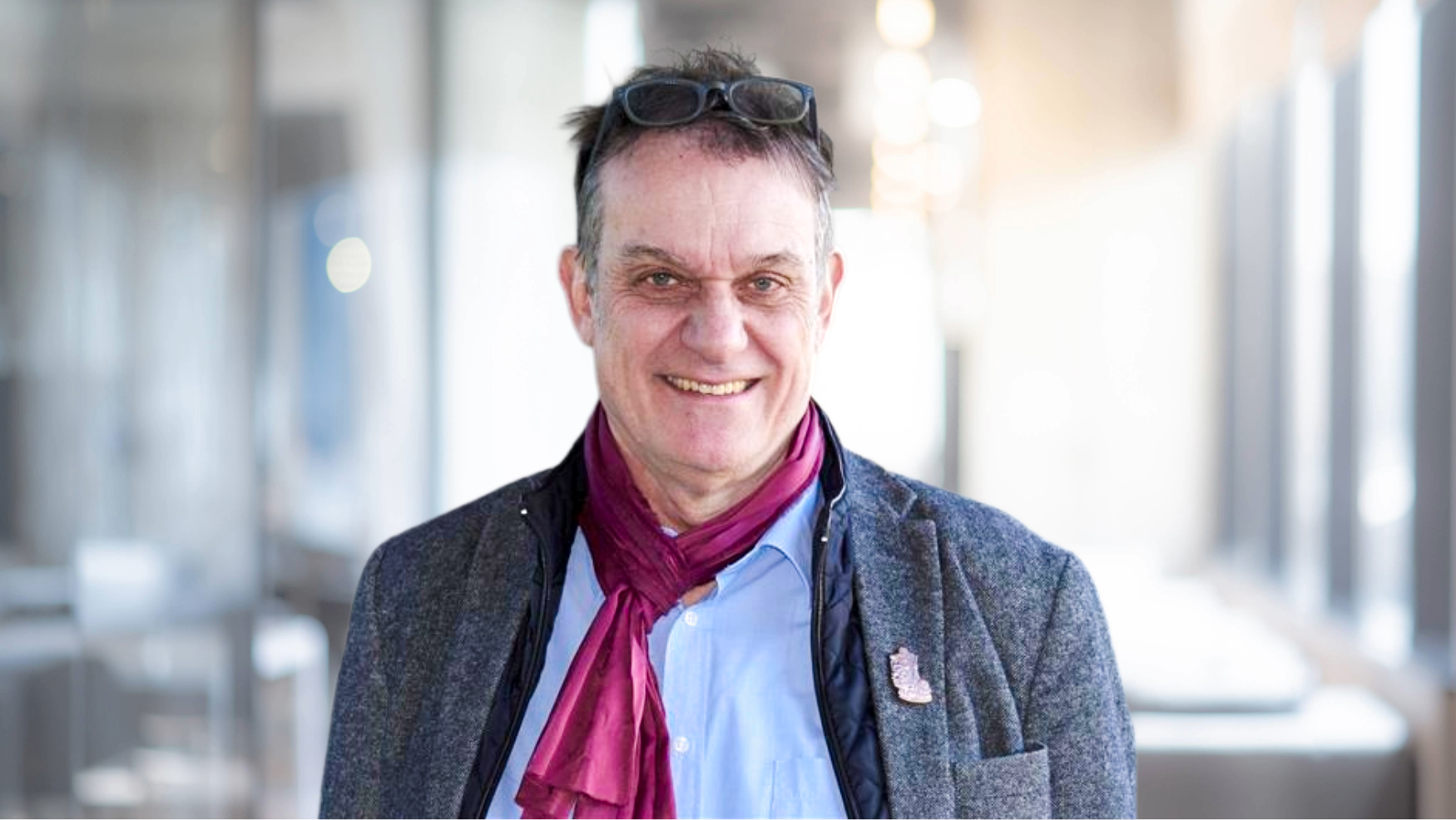 Ein älterer Mann mit grauen Haaren, Brille auf dem Kopf, trägt ein hellblaues Hemd, einen grauen Blazer und eine rosa Halskrause, lächelt freundlich in die Kamera.