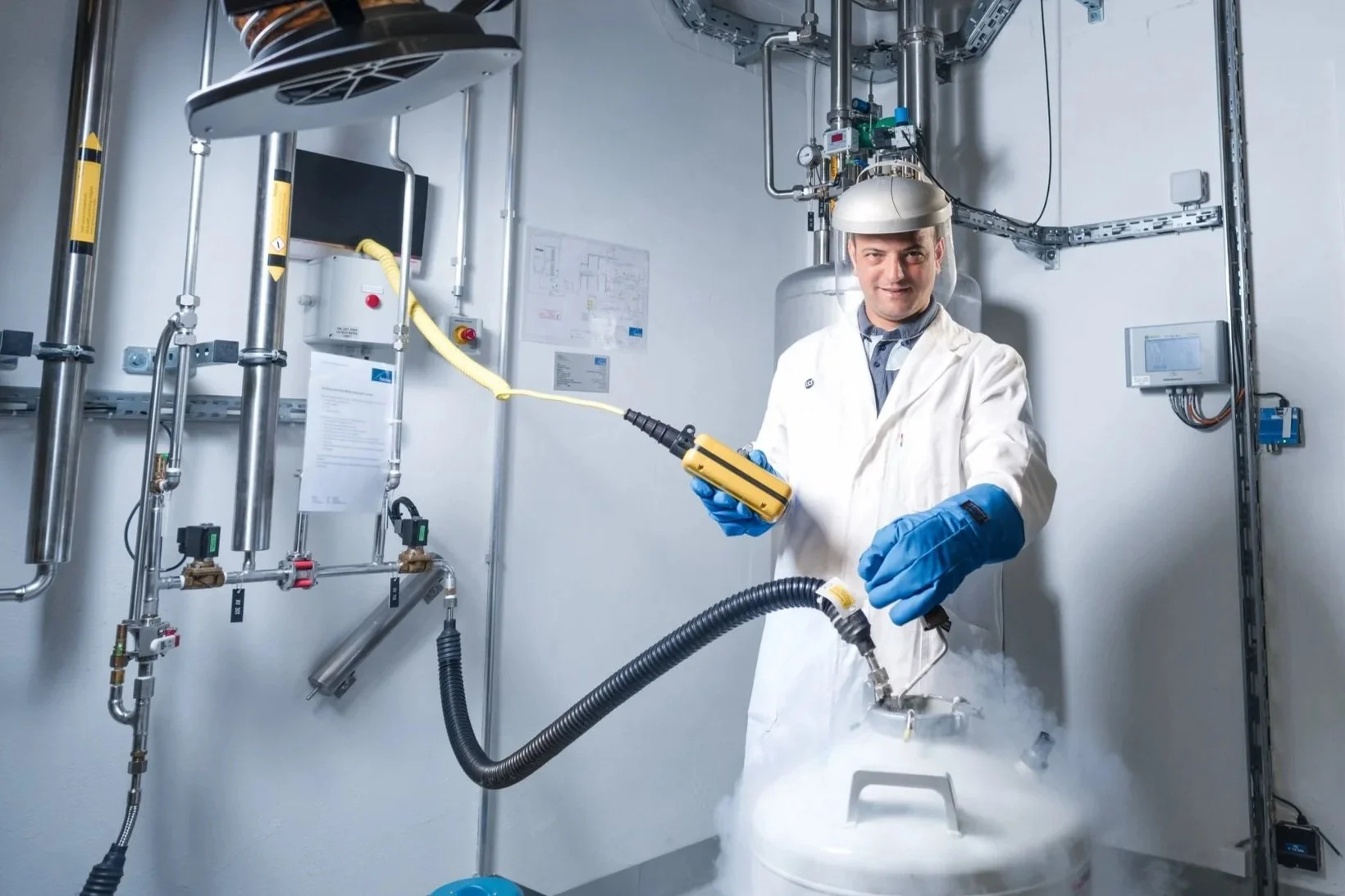 Ein Wissenschaftler trägt eine Schutzkittel, blaue Handschuhe und einen Schutzhelm, während er mit einem technischen Gerät in einem Labor arbeitet, das Dampf oder Rauch erzeugt.