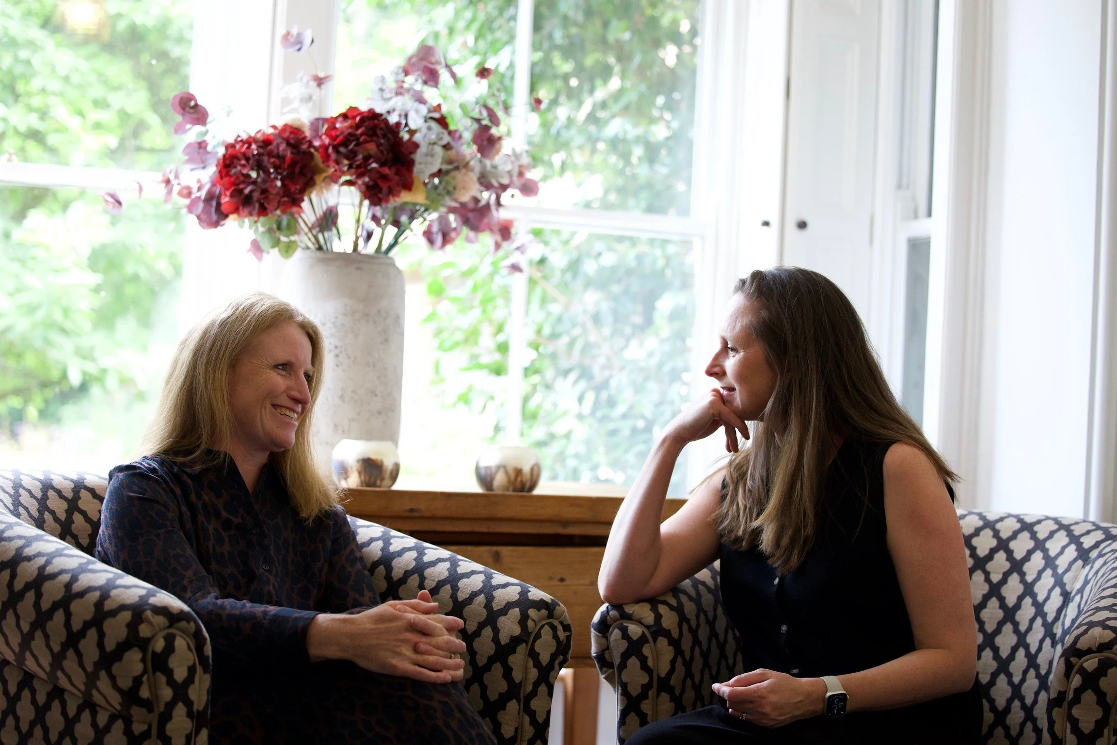Two women sitting on patterned armchairs having a conversation in a bright room with large windows and a vase of red and white flowers in the background.