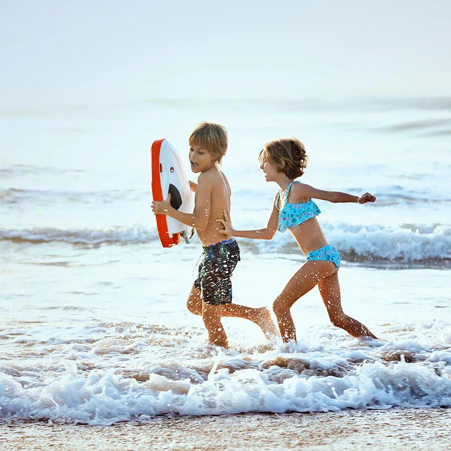 Two children playing in the ocean waves at the beach, one holding a kickboard: Sublue Swii, wearing swimsuit, and the other in a blue swimsuit, with sunlight and calm water in the background.