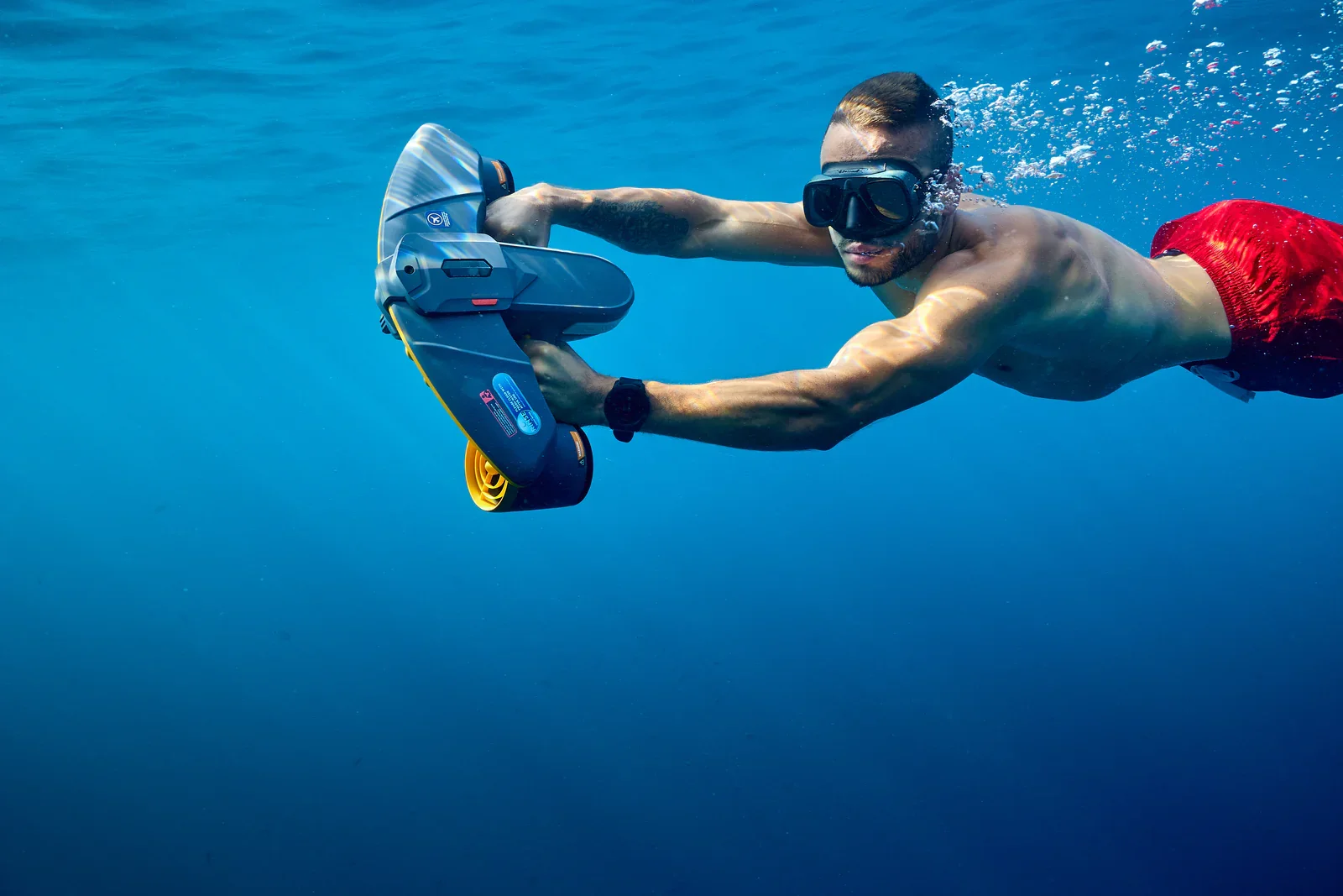 Man swimming underwater with Sublue Navbow+ orange color, wearing goggles and red swim trunks.