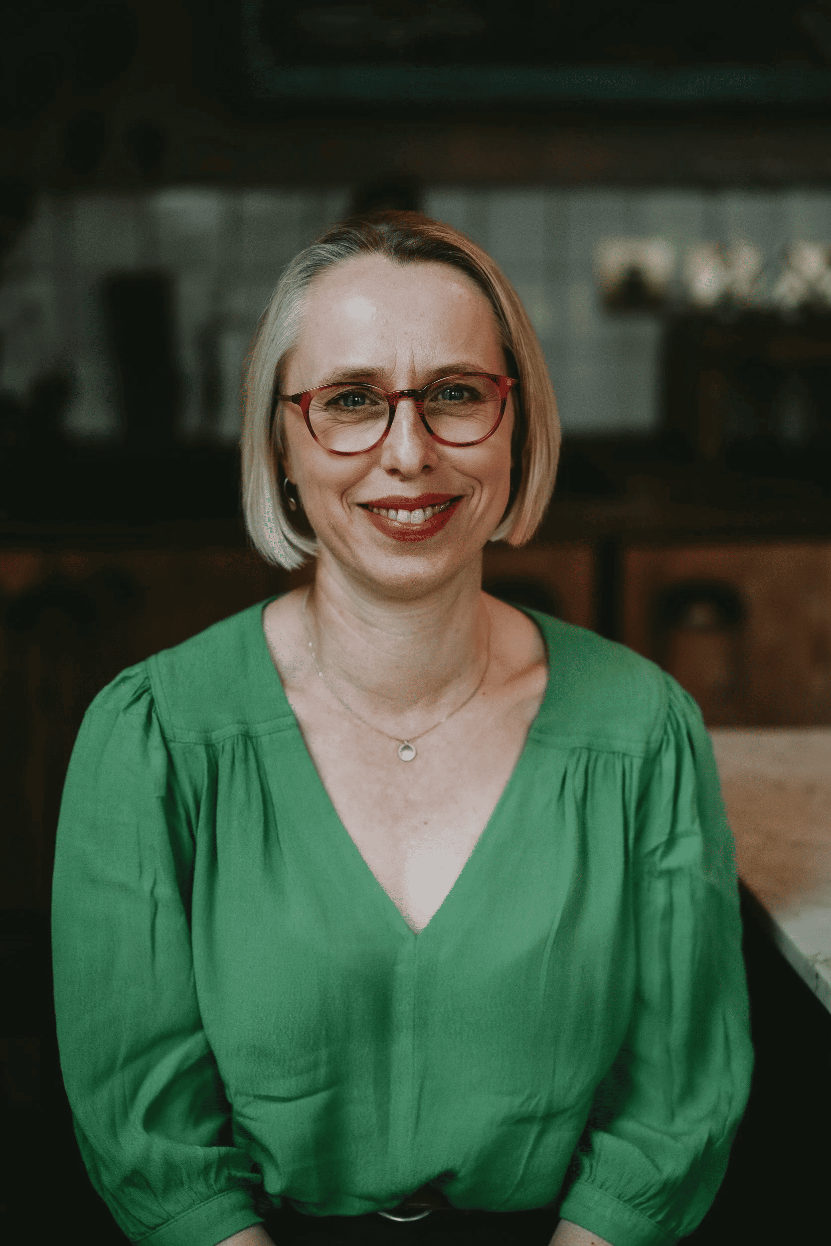 A woman with blond hair, red glasses, and red lipstick sitting at a table, smiling, wearing a green blouse and a delicate necklace in a cozy, rustic setting.