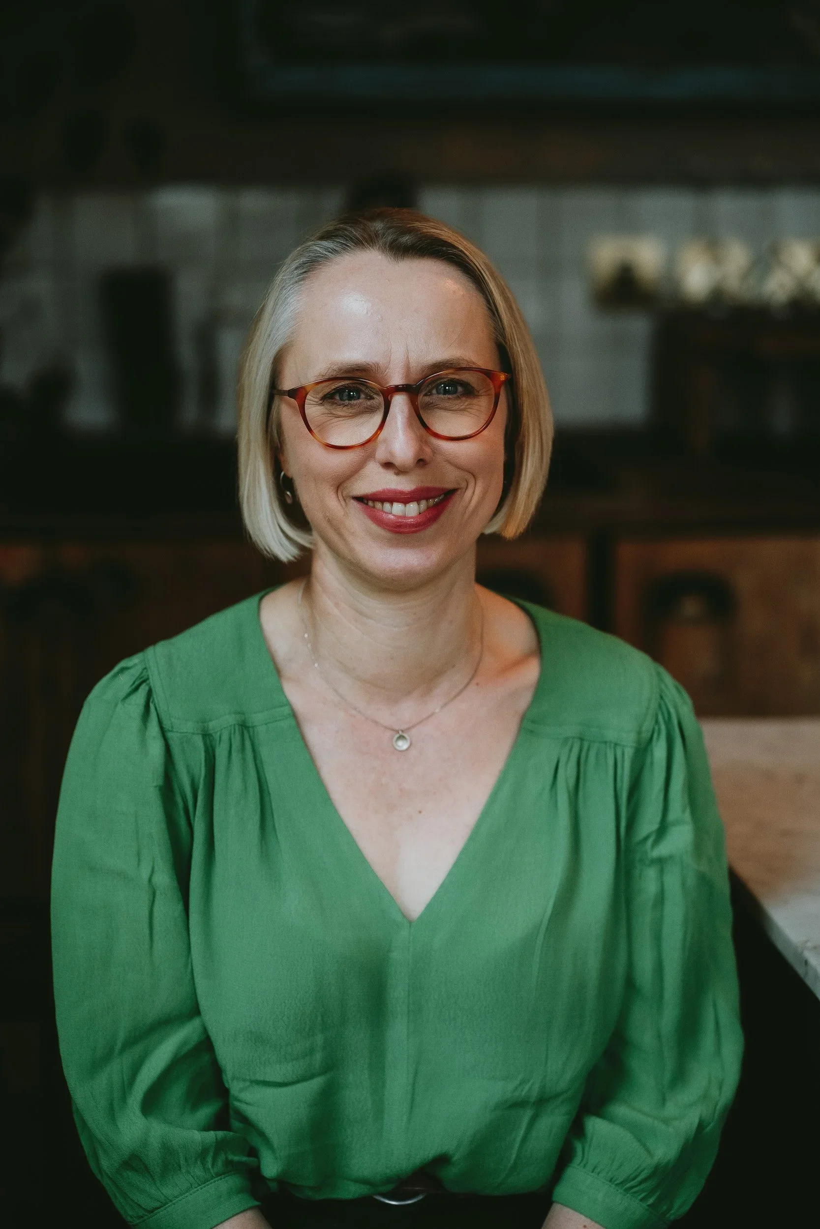 Smiling woman with blonde hair in a bob, wearing glasses and a green blouse, seated in a room with a blurred background.