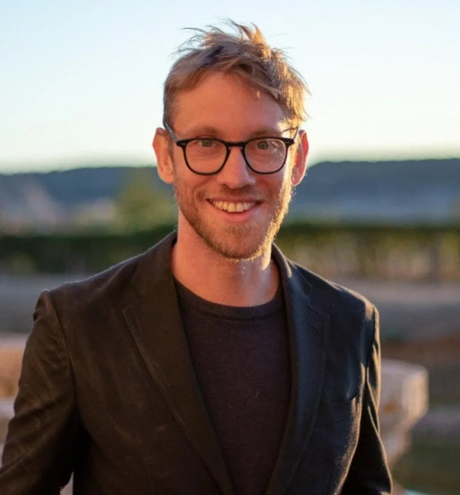 A smiling man with glasses and tousled hair, wearing a black blazer, standing outdoors during sunset with a blurred landscape in the background.