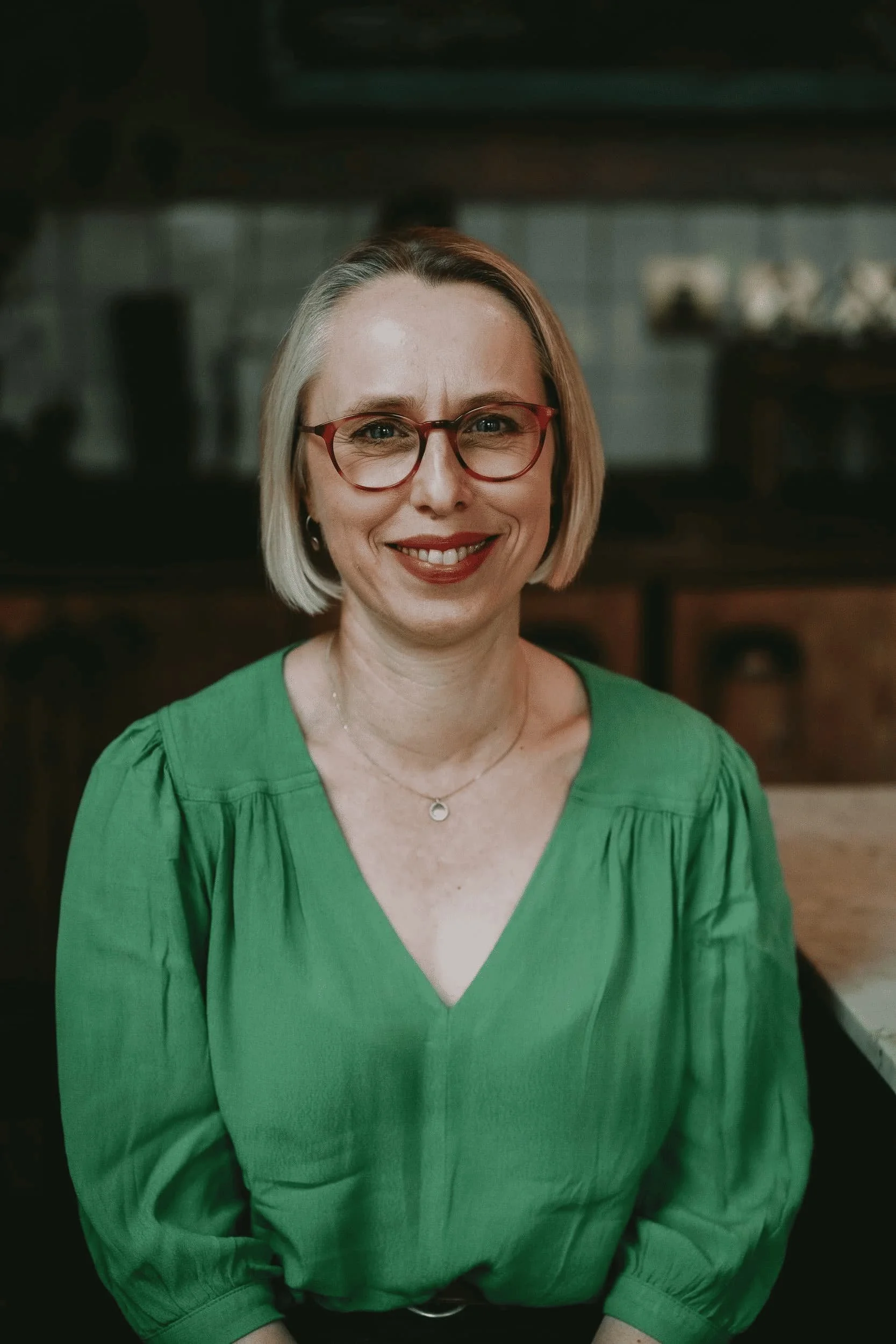 A woman with shoulder-length blonde hair wearing red-rimmed glasses, a green blouse with puffed sleeves, smiling in an indoor setting with blurred background.