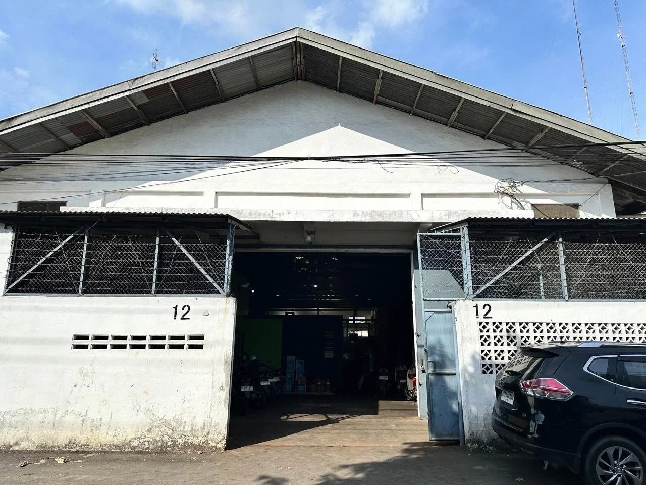Front view of a white industrial or warehouse building with the number 12 on the wall and gate, black car parked outside, open entrance showing motorcycles inside, with a clear blue sky overhead.