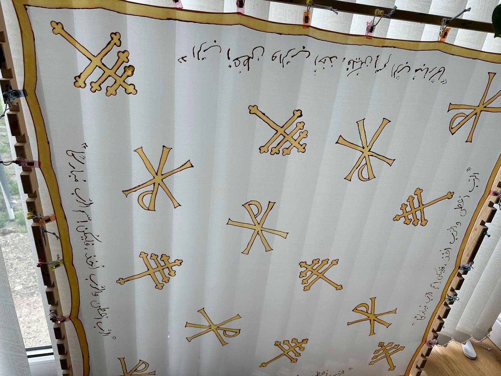 A religious cloth with several gold-colored crosses and Arabic script along the edges, displayed hanging in a room with a window to the side.