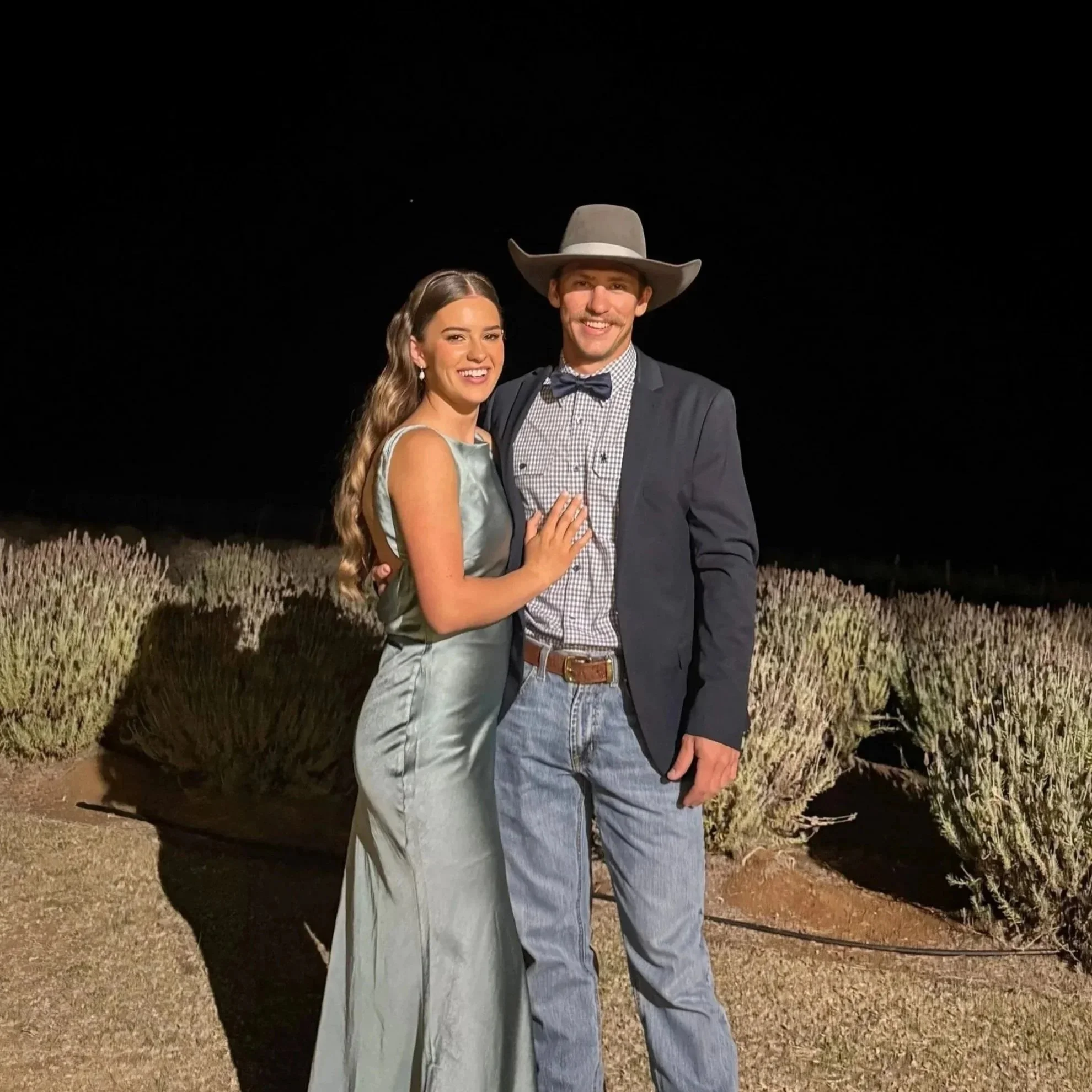A young woman and a young man posing together outdoors at night. The woman is wearing a sleeveless, shiny silver dress, and the man is wearing a dark blazer, a checked shirt, jeans, a belt, and a cowboy hat. They are smiling and the background features bushes or plants under a dark sky.