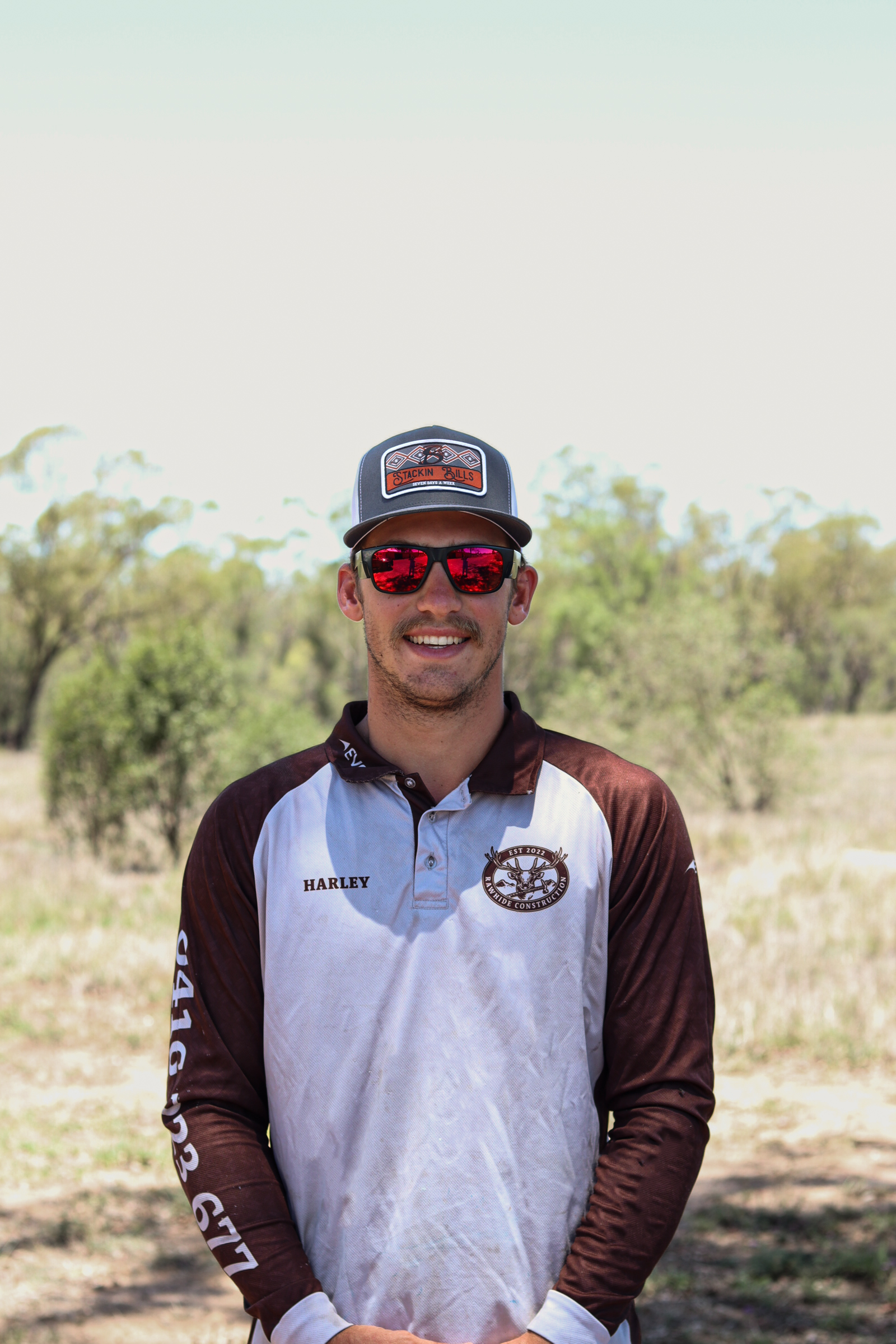 A smiling man wearing red sunglasses, a gray and brown sports jersey, a gray cap with a logo, standing outdoors in a grassy area with trees in the background.