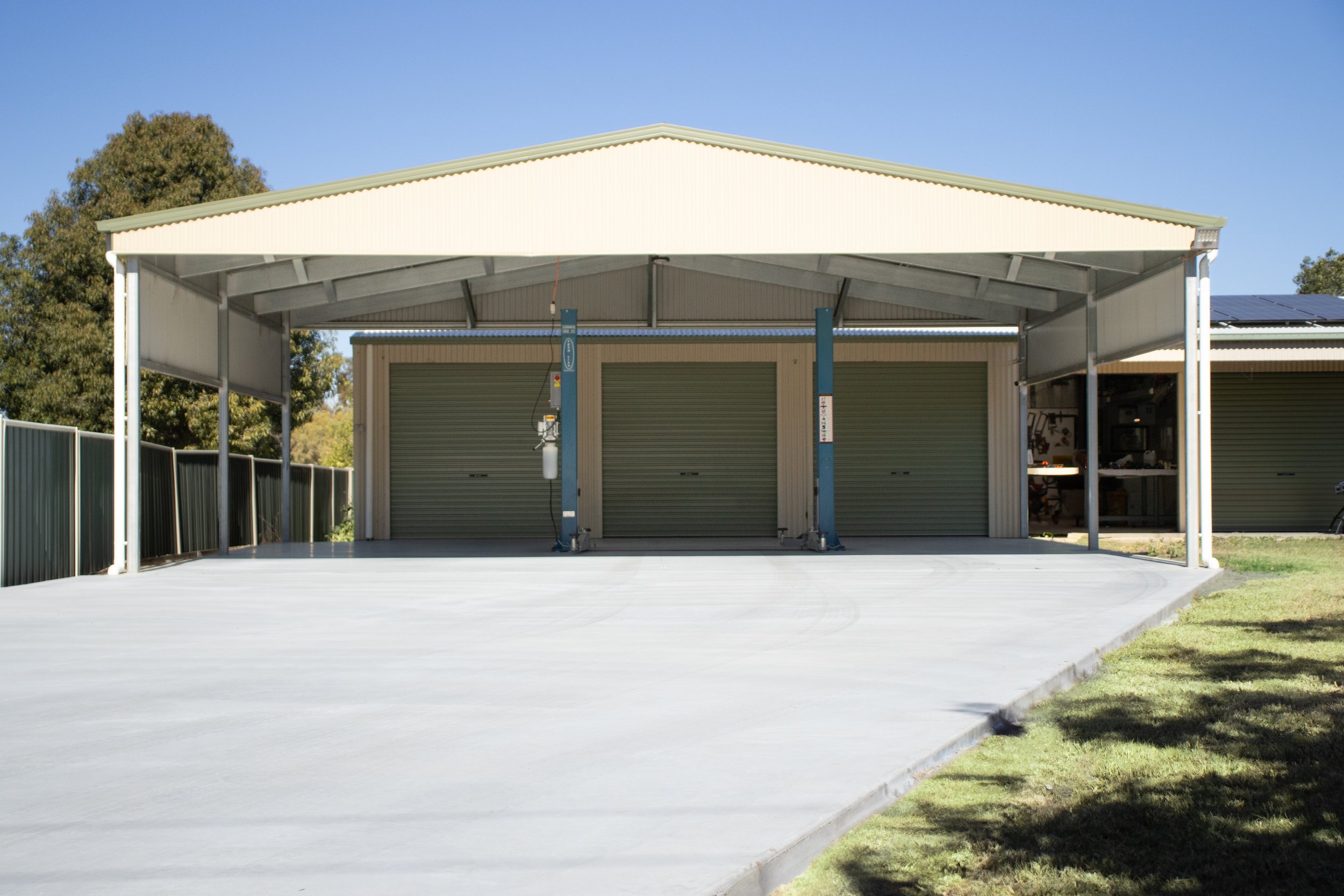 Empty carport with three garage doors, concrete driveway, and a metal fence on the left, with trees and a blue sky in the background.