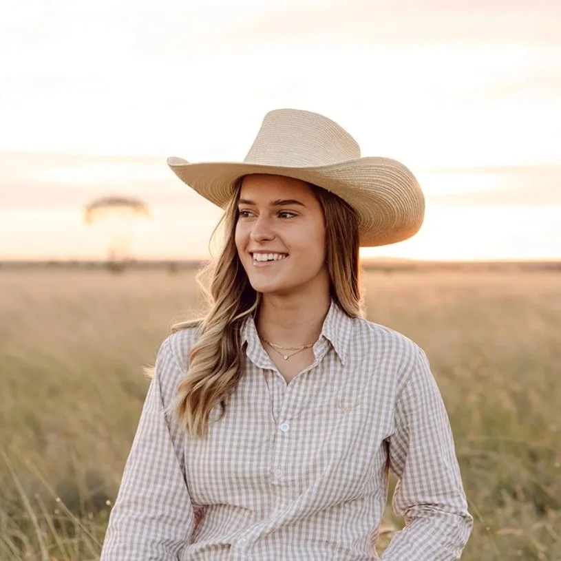Young woman in a wide-brimmed hat, plaid shirt, outdoors during sunset, smiling