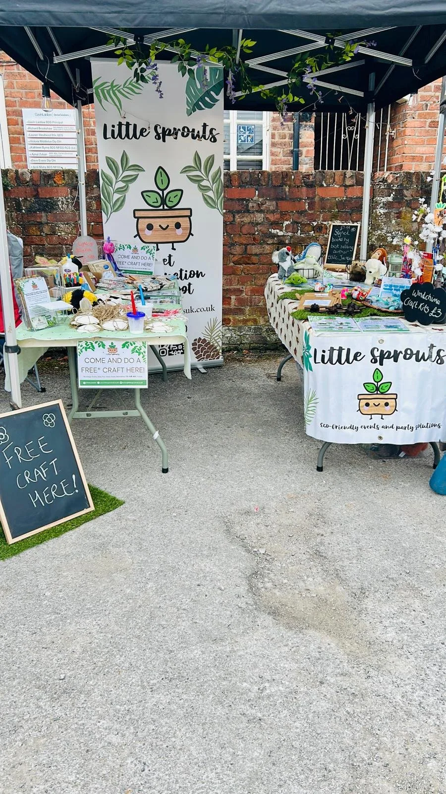 Outdoor craft stall with tables displaying various handmade items, sign advertising free craft activities, and a banner with a cute sprout character for 'Little Sprouts' eco-friendly events and party solutions, set against a brick wall background.