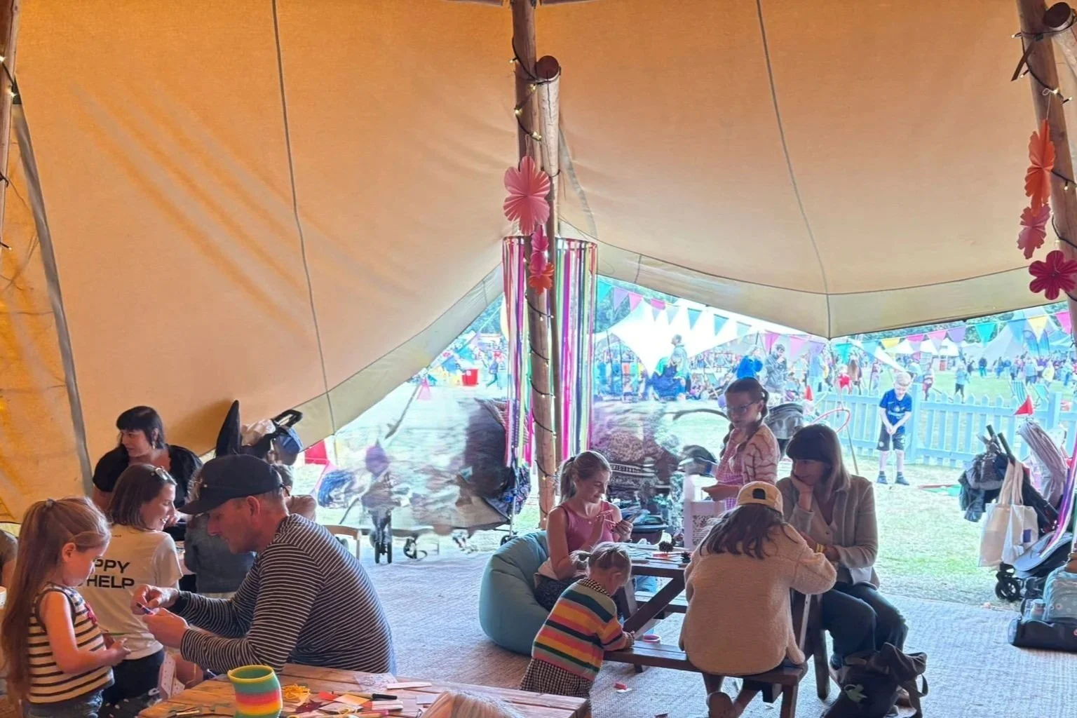 Children and adults gathered inside a large beige fabric tent with colorful decorations, including pink and purple flowers and streamers, at an outdoor festival or fair.