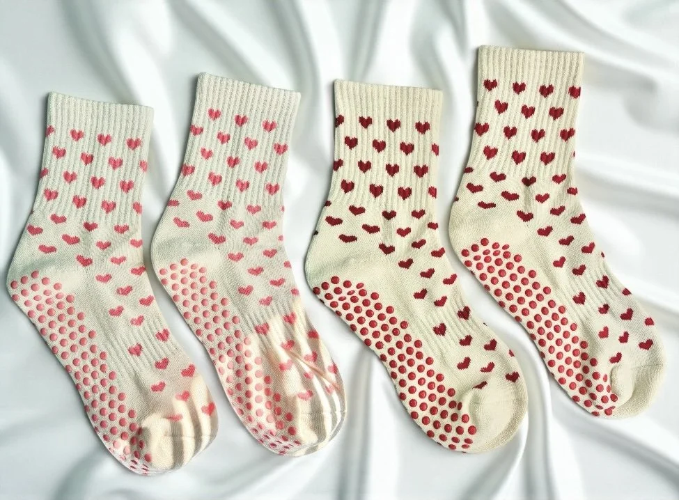 Four pairs of cream-colored socks with pink and red heart patterns and red polka dots on the soles, laid out on a white silk fabric background.