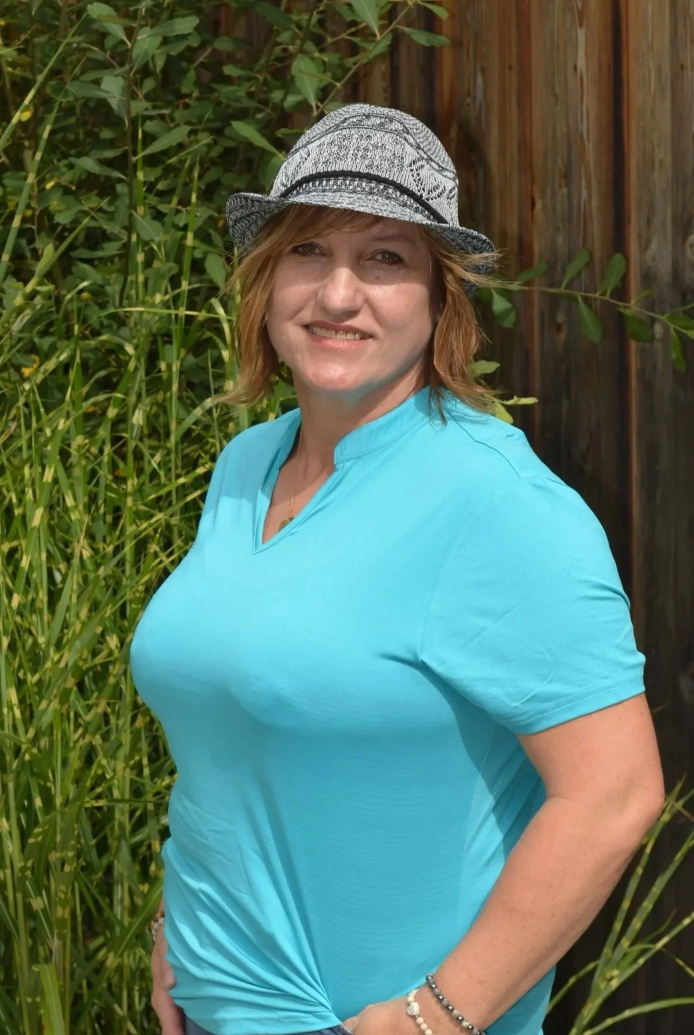 Eine Frau mit braunen Haaren trägt einen Strohhut, eine hellblaue Bluse und mehrere Armbänder, steht vor grünen Pflanzen und einem Holzzaun.