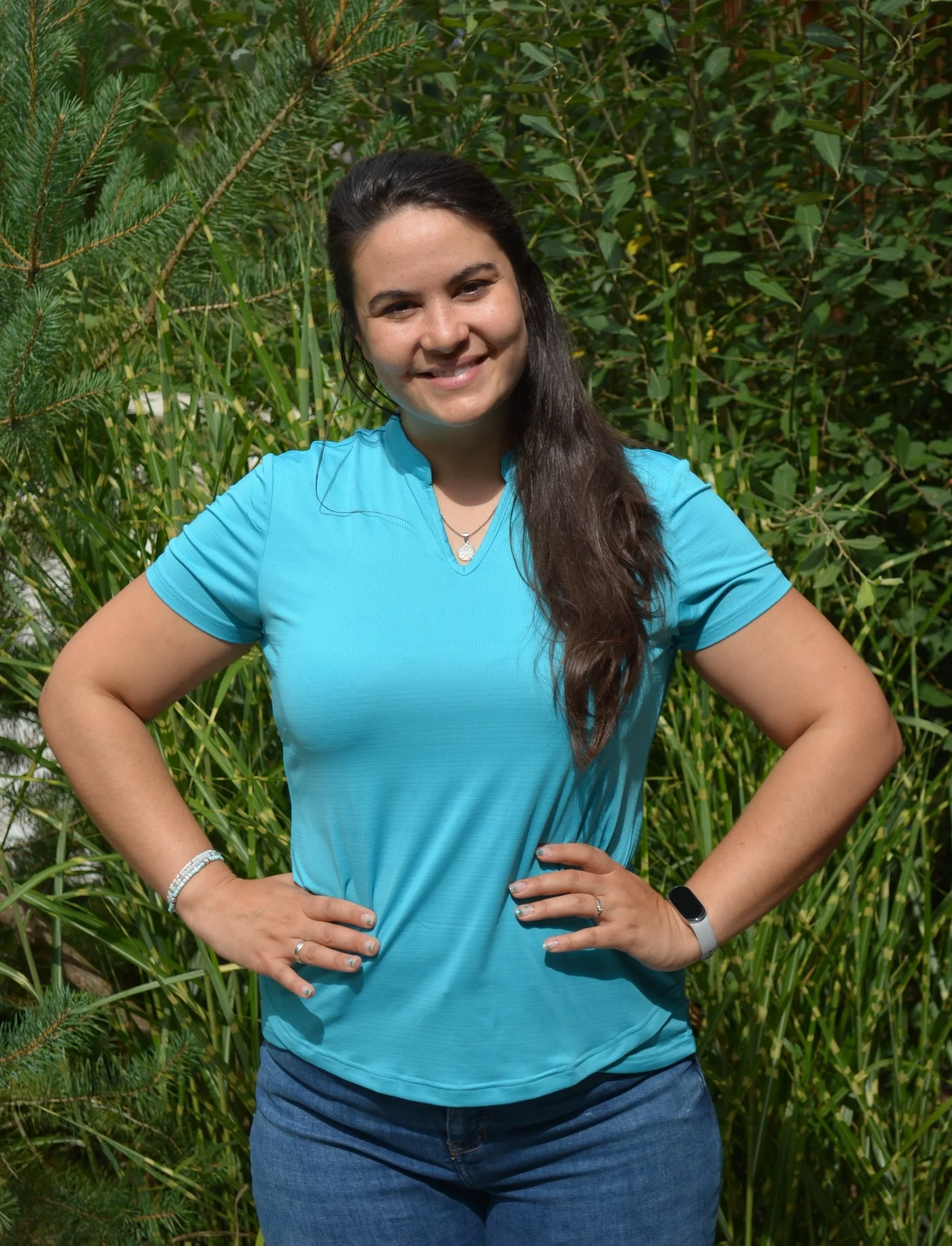 Junge Frau mit dunkelbraunen Haaren in blauer Sportjacke vor grünen Sträuchern, lächelnd, mit Hand auf Hüfte, trägt Armband, Ring, und Smartwatch.