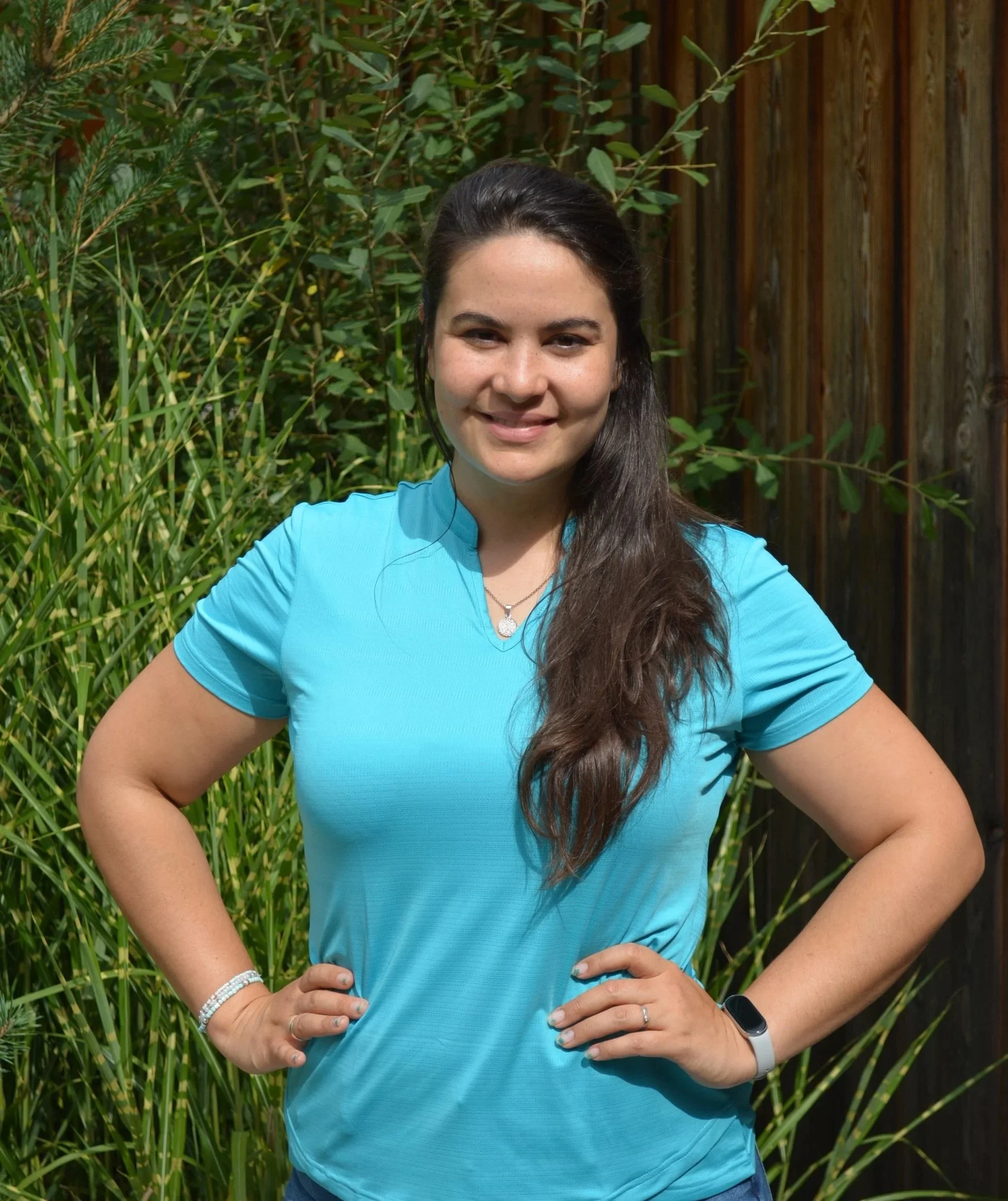 Eine junge Frau steht im Garten vor grünen Pflanzen und einem Holzzaun, trägt ein hellblaues T-Shirt, eine Halskette, einen Ring, ein Armband und eine Smartwatch, lächelt und hat die Hände auf den Hüften.