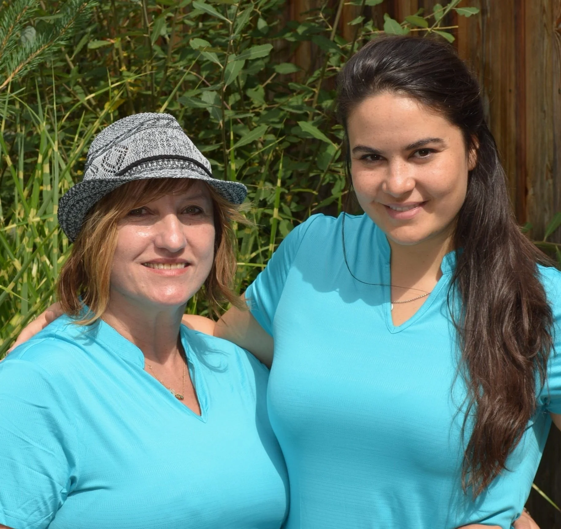 Zwei Frauen in blauen Shirts stehen vor grünen Pflanzen und lachen zusammen.