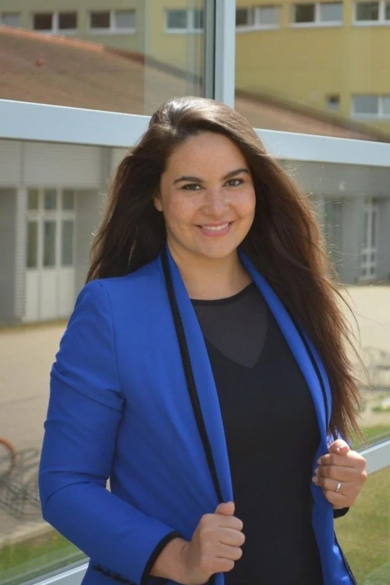 Eine lachende Frau mit brünettem, langen Haar, die eine blaue Jacke trägt, steht vor einem Fenster mit moderner Gebäude im Hintergrund.