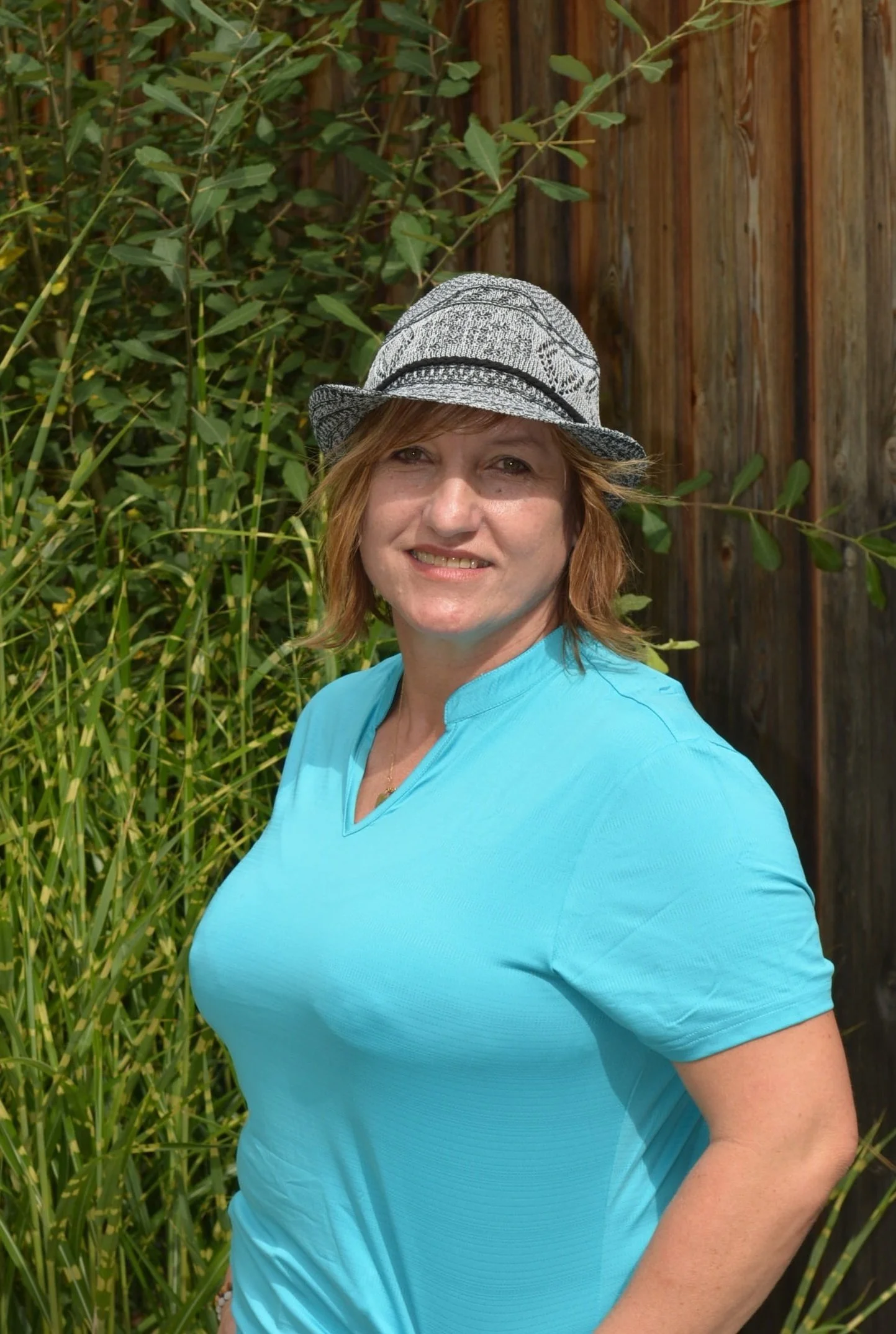 Eine Frau trägt einen grauen Hut und ein hellblaues T-Shirt, steht vor einem Holzzaun und grünen Pflanzen im Hintergrund.