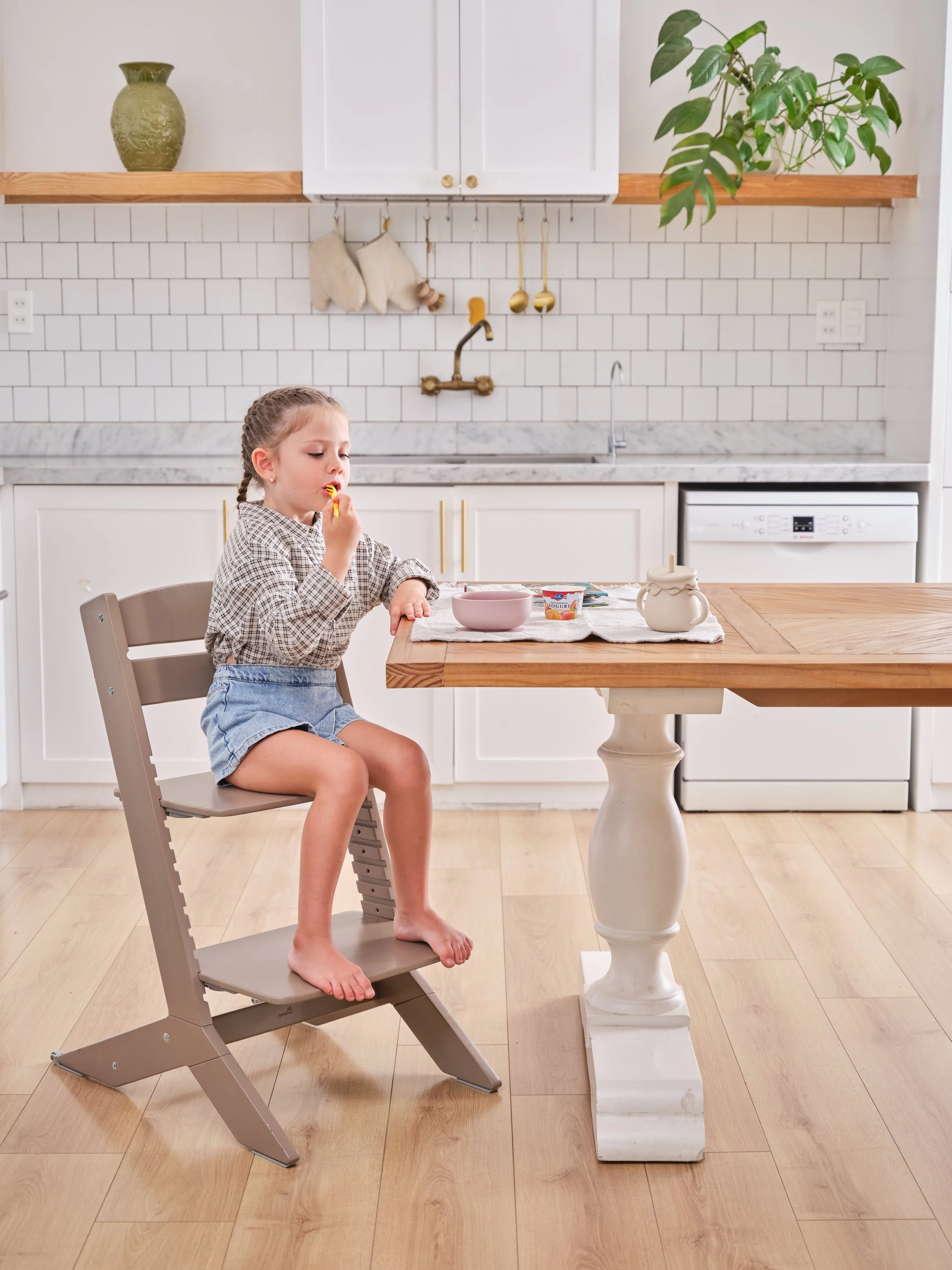 A young girl sitting on a high chair at a kitchen table, eating yogurt with a spoon. The kitchen has white cabinets, a white subway tile backsplash, and a wooden countertop. There are a few small cups and a teapot on the table.