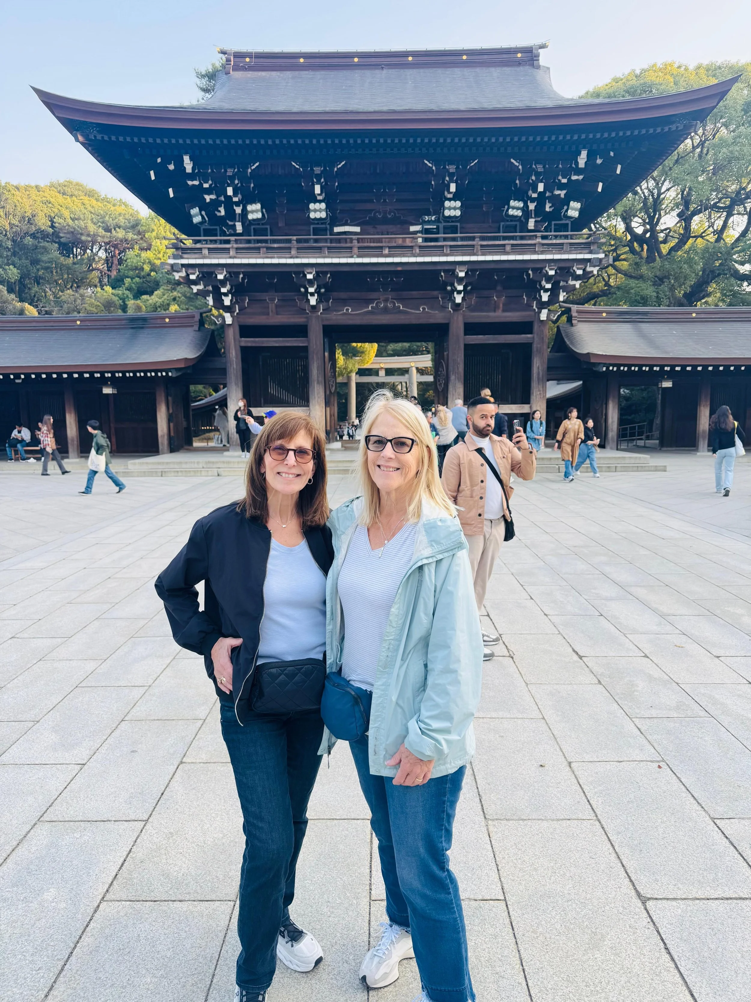 Two women in casual clothing and glasses posing and smiling for a photo in front of a traditional Japanese temple gate. There are other visitors walking in the background.