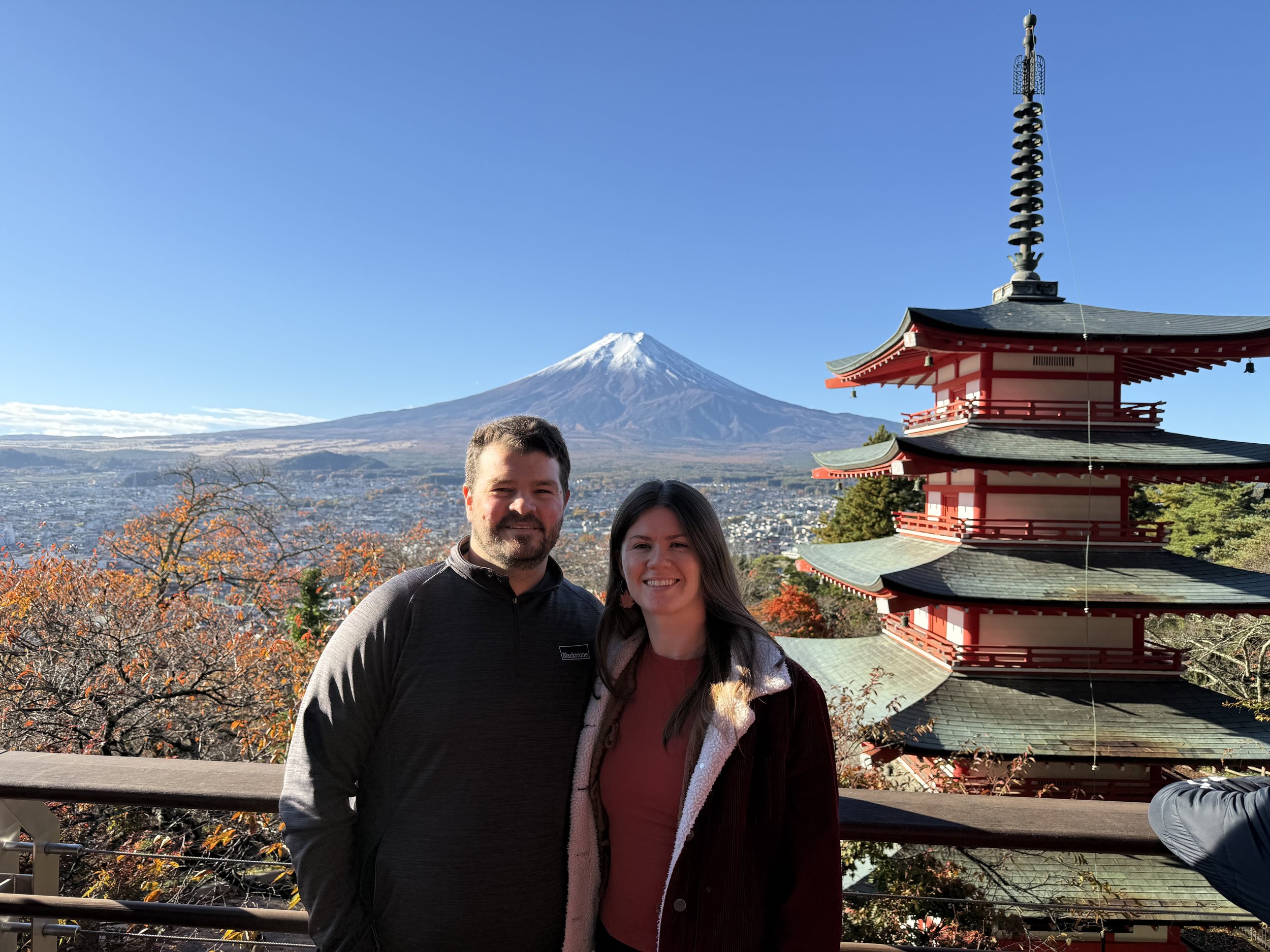 A man and woman standing together outdoors with Mount Fuji in the background, a traditional Japanese pagoda on the right, and autumn trees in the foreground.