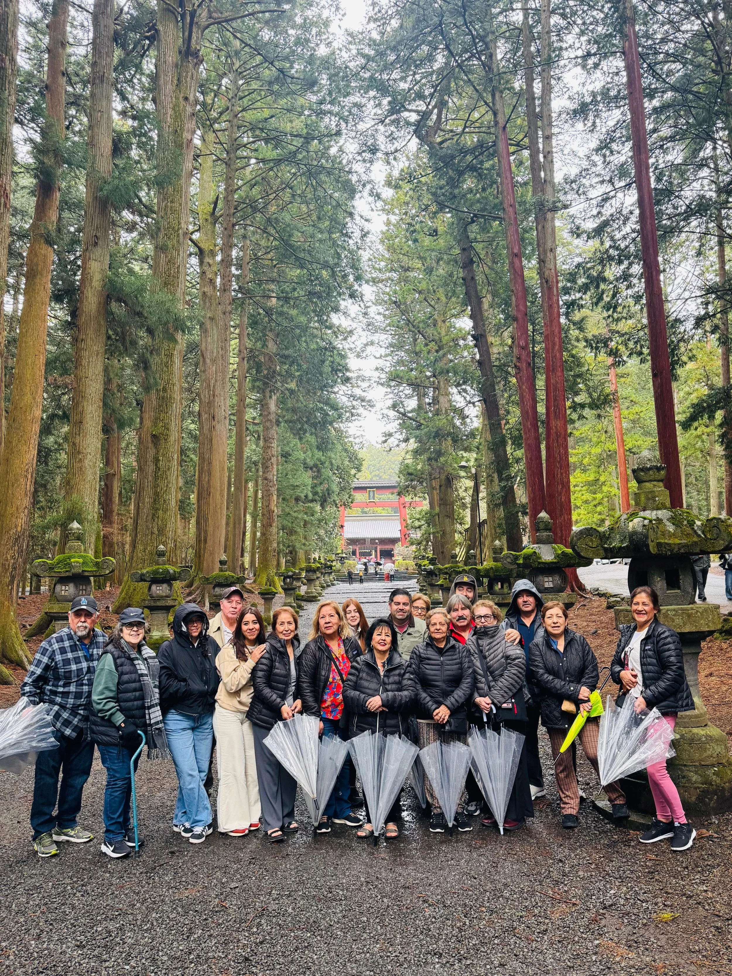 Group of people standing in a forested area with tall trees, moss-covered stone lanterns, and a traditional Japanese shrine in the background.