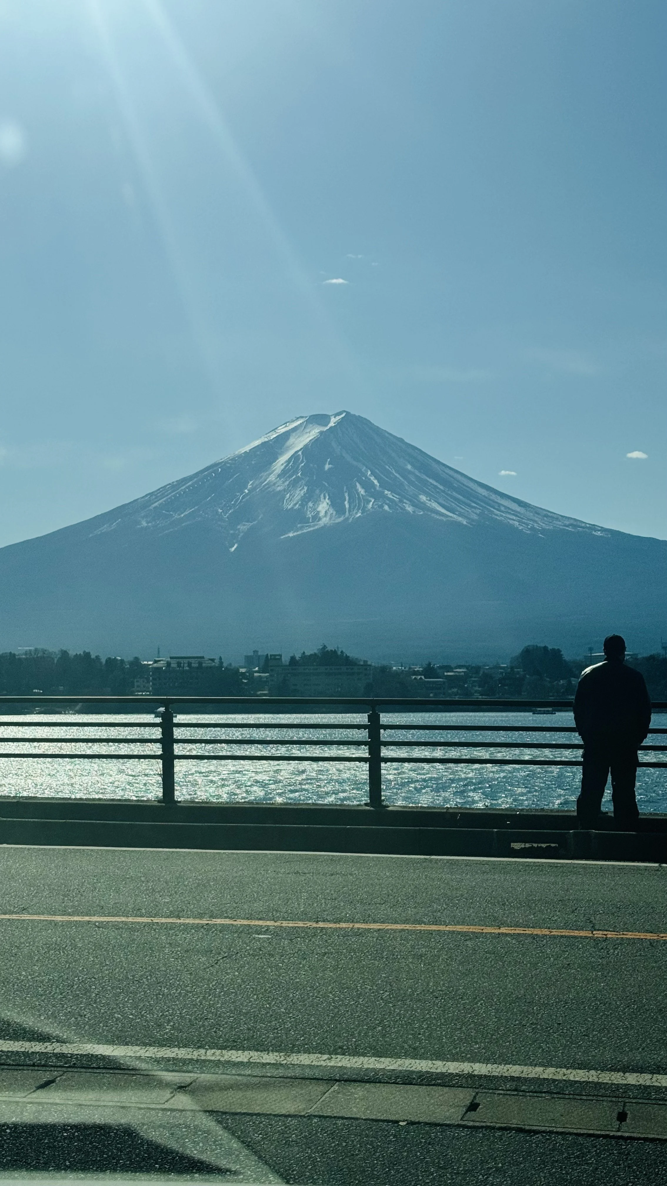 View of Mount Fuji with snow on top, seen across a body of water, with a person standing by a fence on the roadside in the foreground.