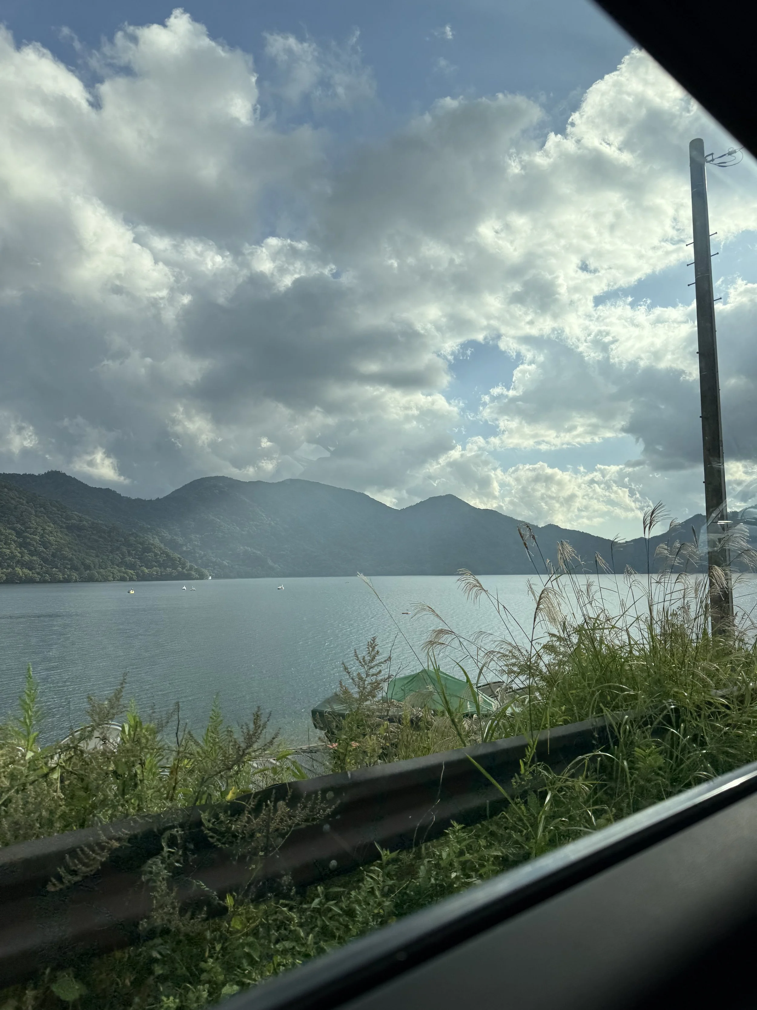 View of a lake surrounded by mountains with a cloudy sky, seen from inside a vehicle with a window and a roadside guardrail.