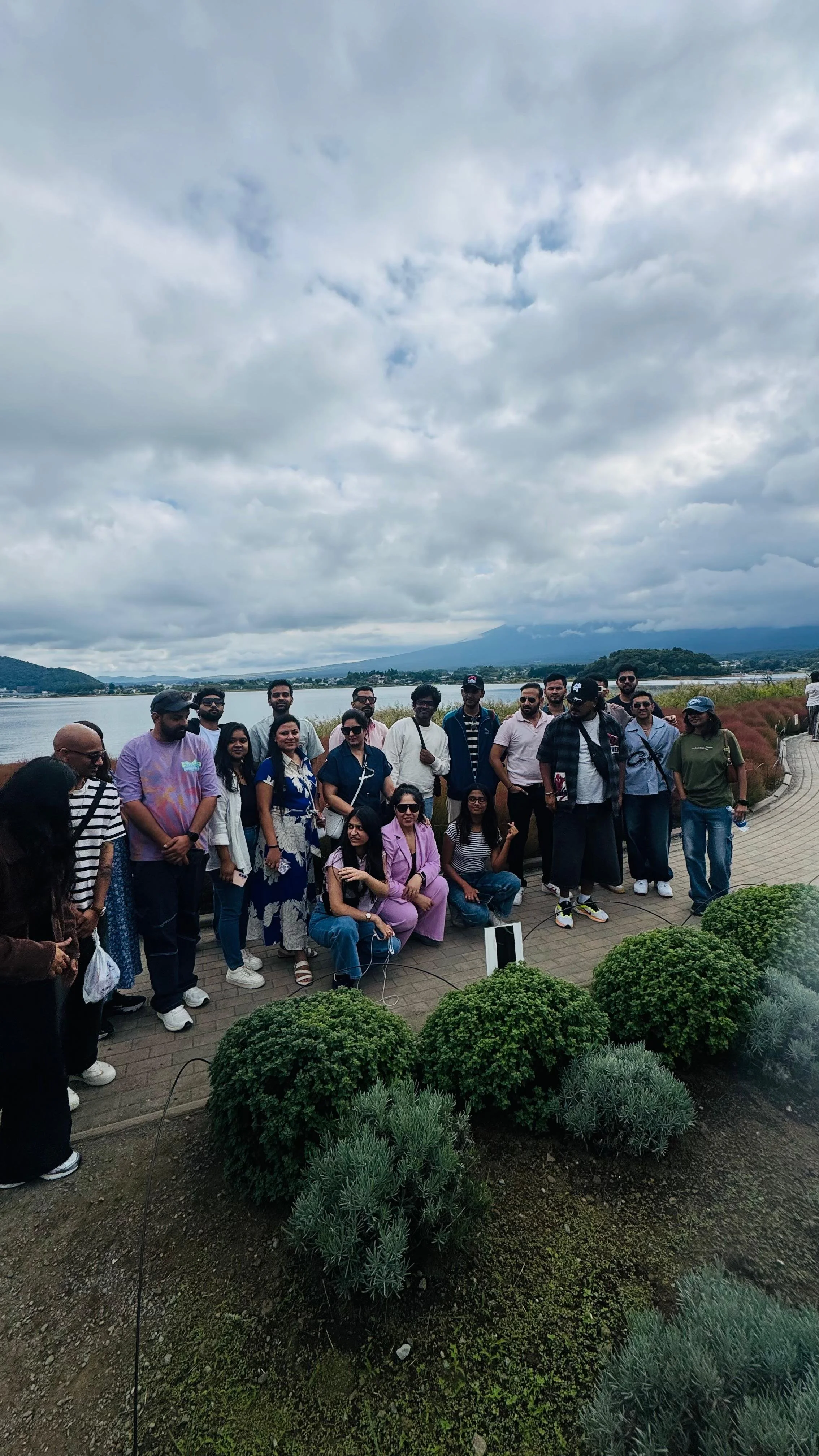 Group of people posing outdoors with a lake and mountains in the background under cloudy sky.