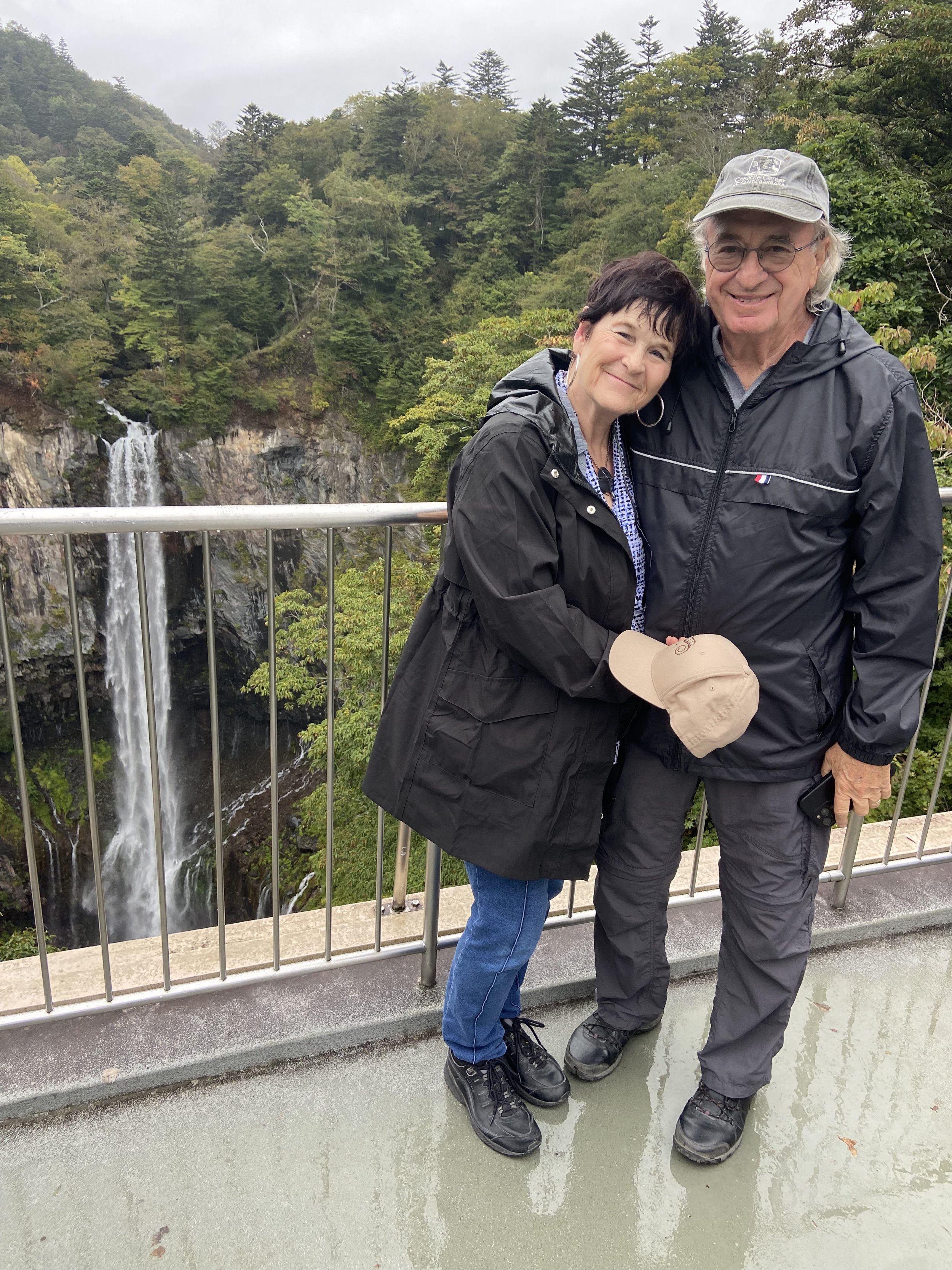A smiling woman and man in rain jackets standing close together on a viewing platform with a waterfall and forest background, holding a beige cap and a phone.