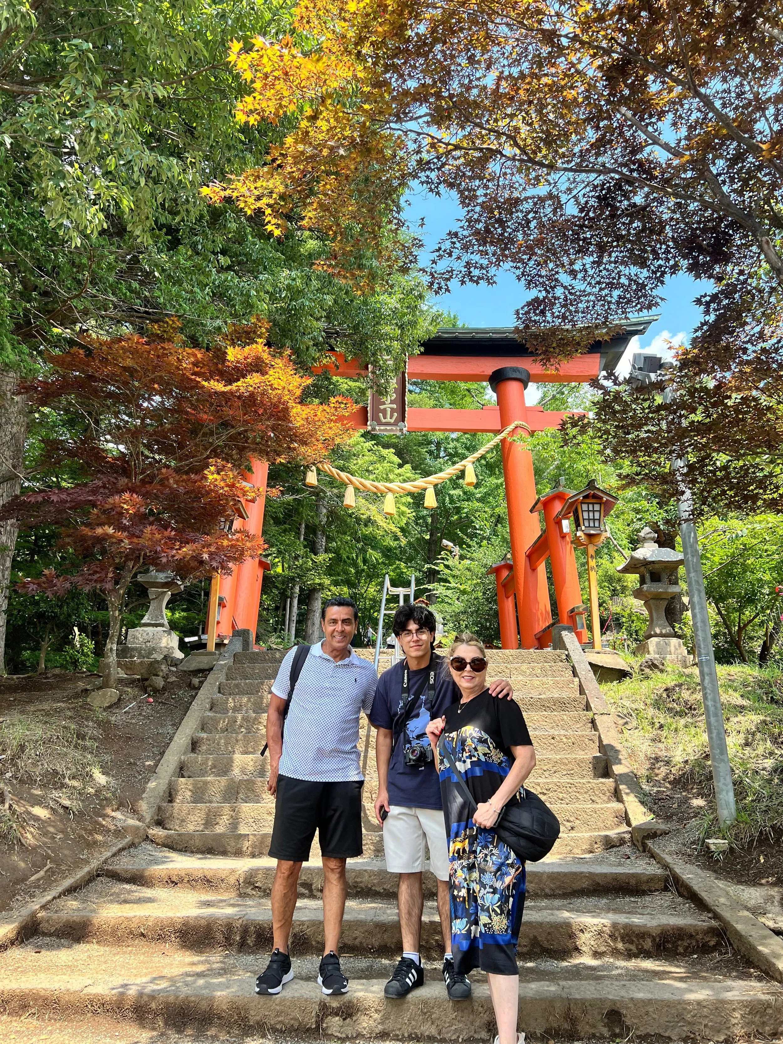 Three people standing on stone steps in front of a red torii gate in a forested area, with trees and traditional lanterns around it.