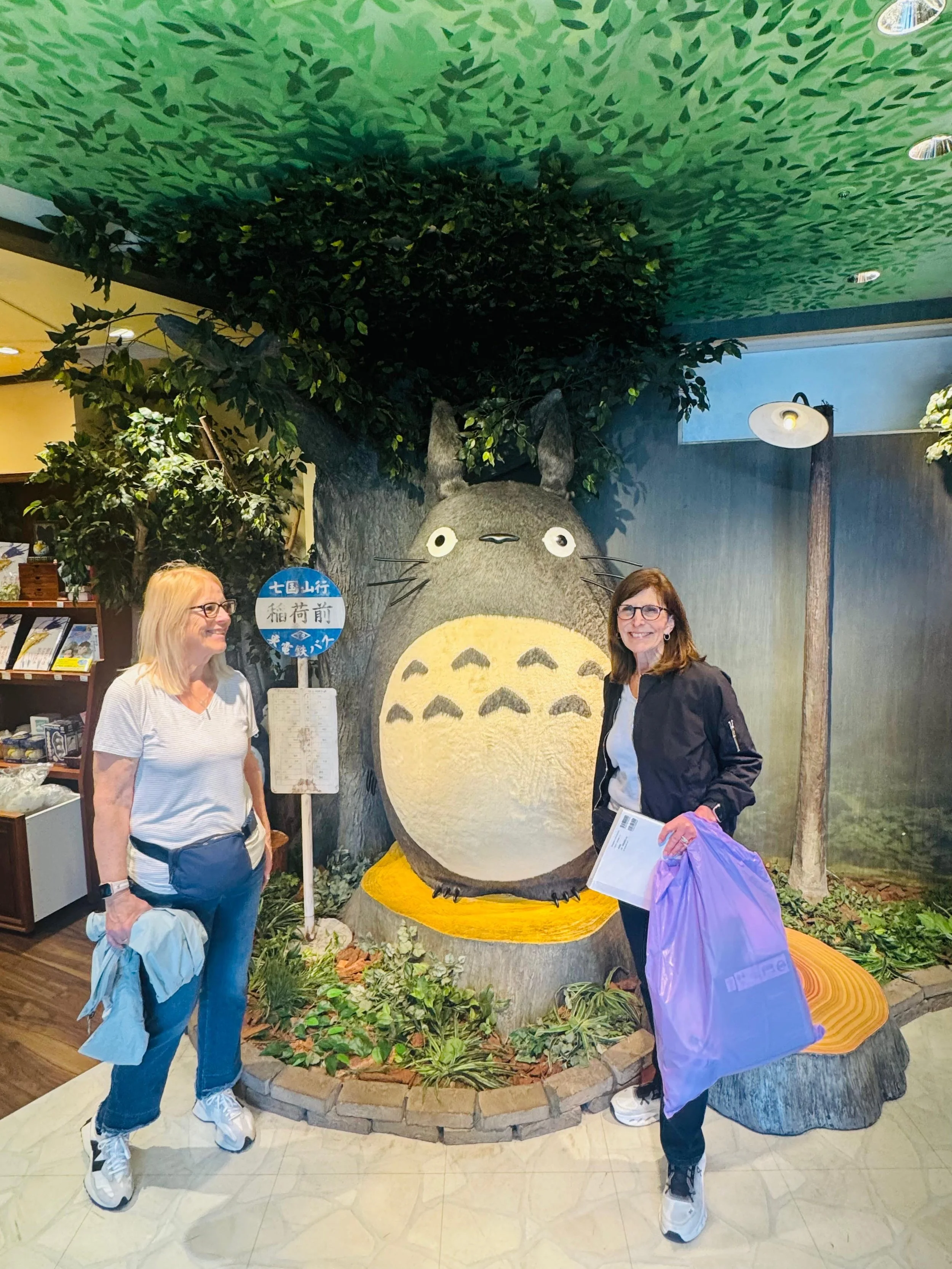 Two women stand beside a large Totoro sculpture made of wood and painted to resemble the character from the movie, with a tree and green forest background.