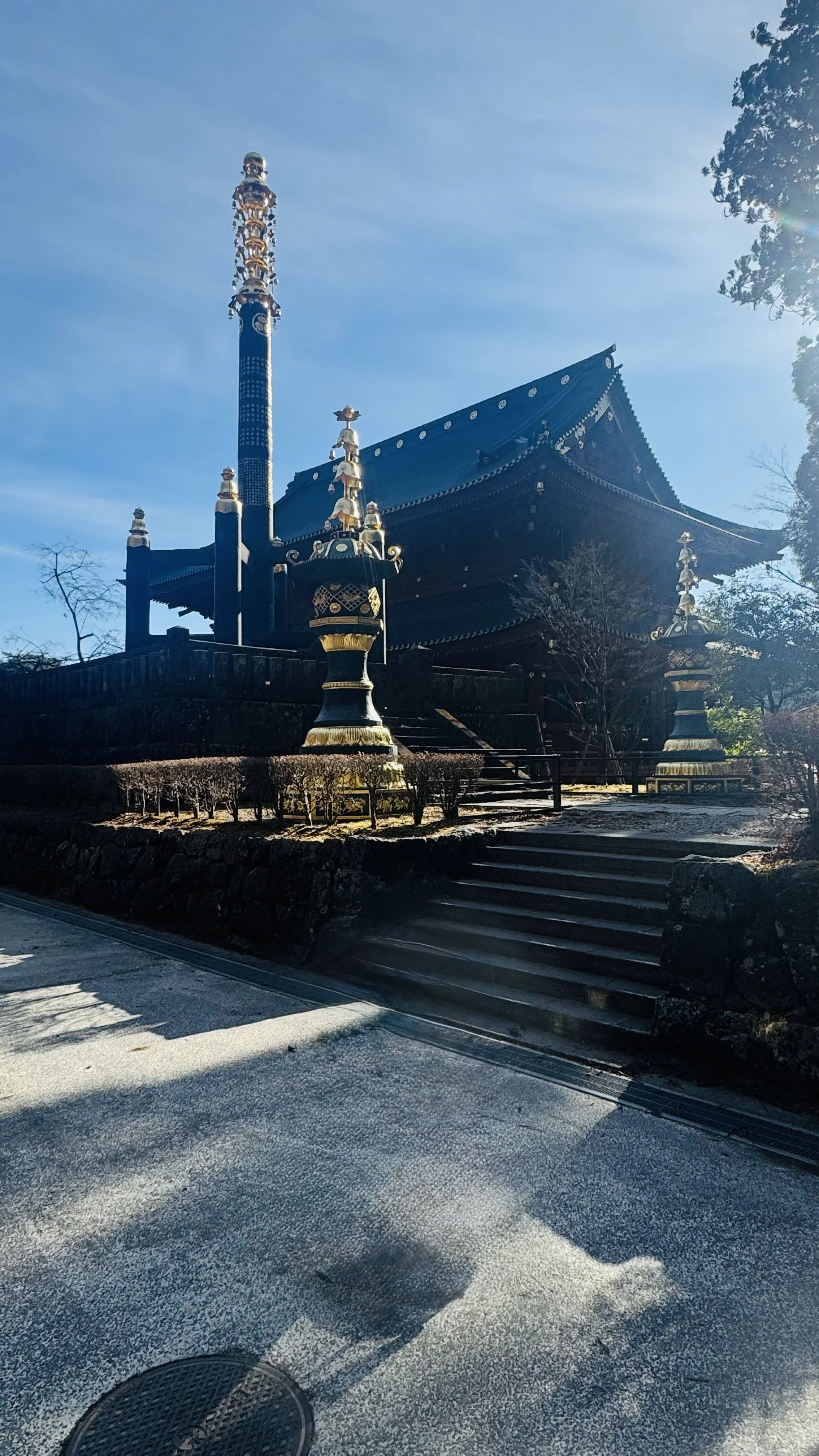 Traditional Japanese temple with black and gold decorations, situated on a small hill with steps and surrounded by trees, under a clear blue sky.