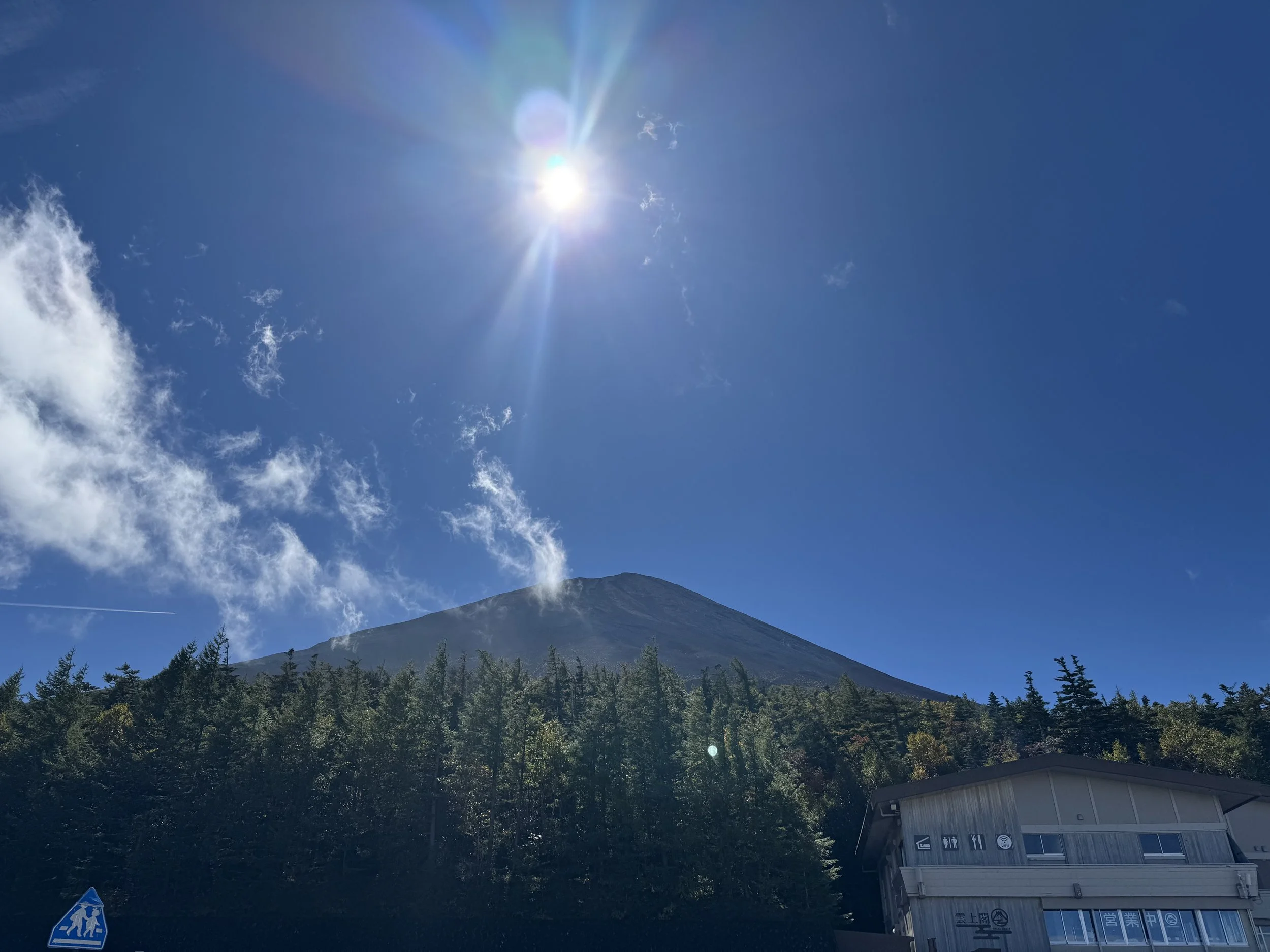 A mountain with a clear blue sky, the sun shining brightly, and some clouds and smoke around the mountain summit. There are trees and a building at the base of the mountain.