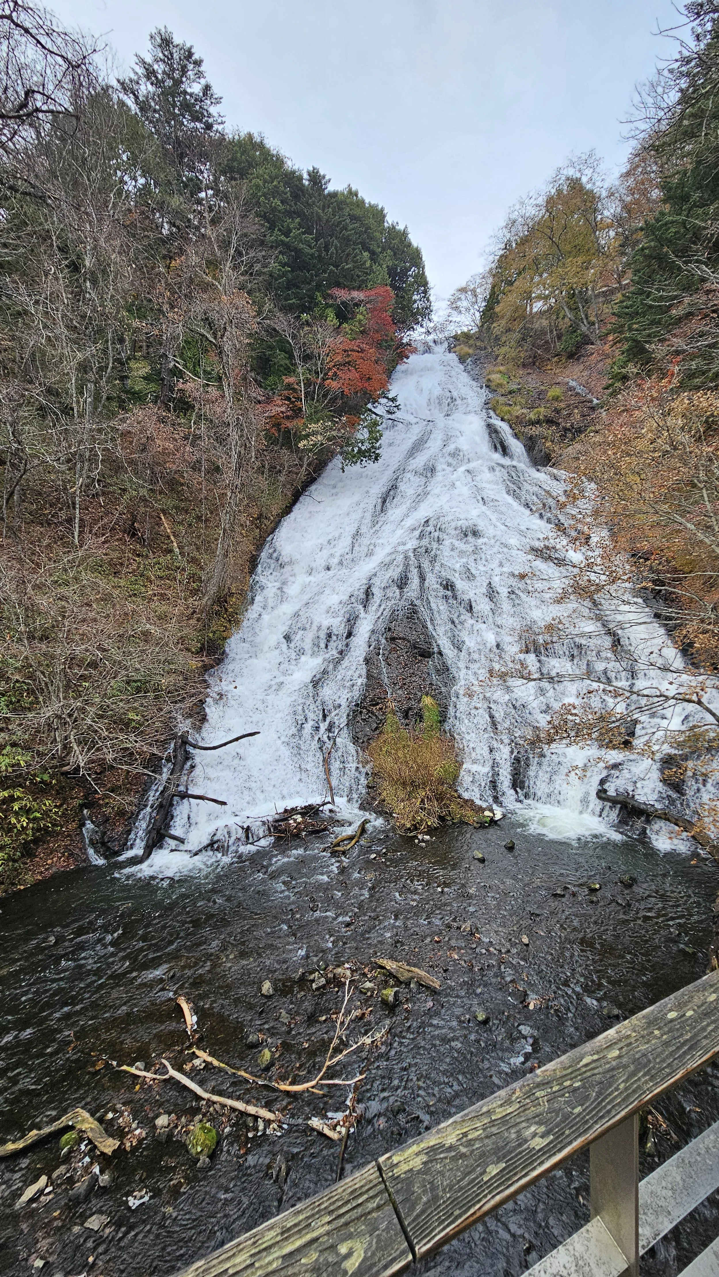 A waterfall flowing down a rocky hillside surrounded by trees with autumn foliage, viewed from a wooden bridge.