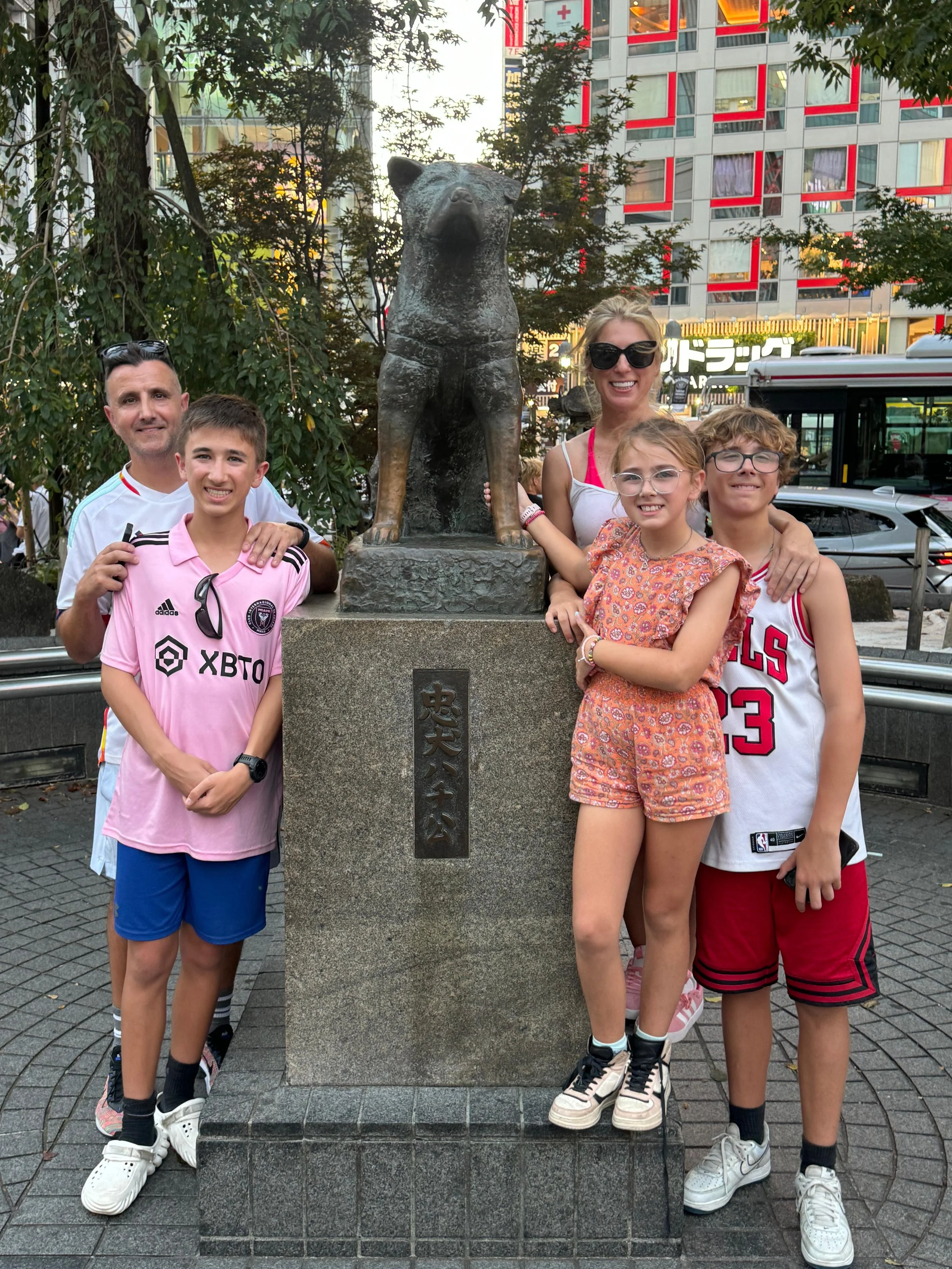 A family of six, including two adults and four children, are posing together outdoors near a bronze statue of a dog on a stone pedestal. The setting appears to be in an urban area with buildings, trees, and a bus in the background.