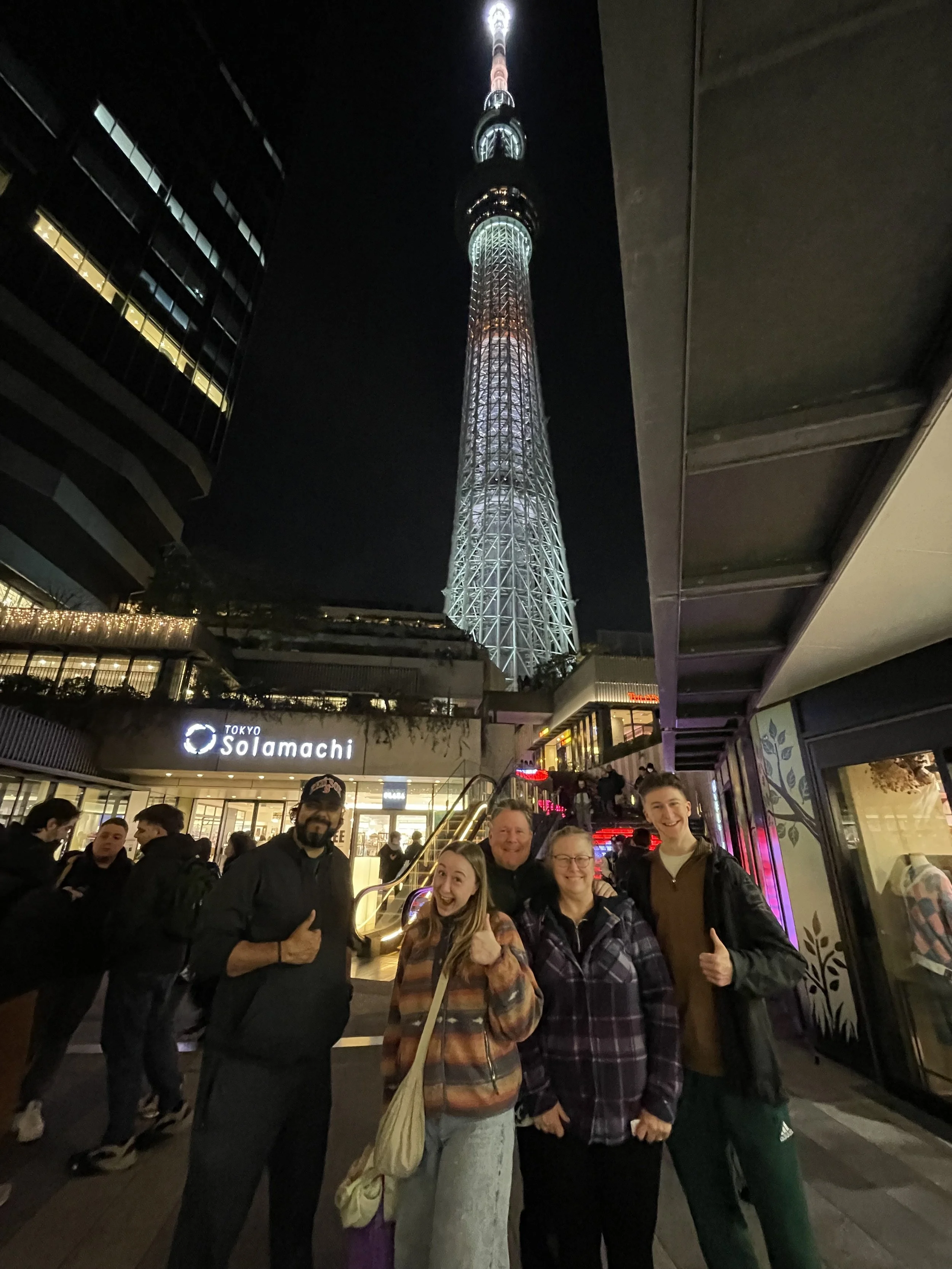 A group of five people smiling and posing for a photo in front of Tokyo Skytree at night, with the Tokyo Solamachi shopping complex in the background.