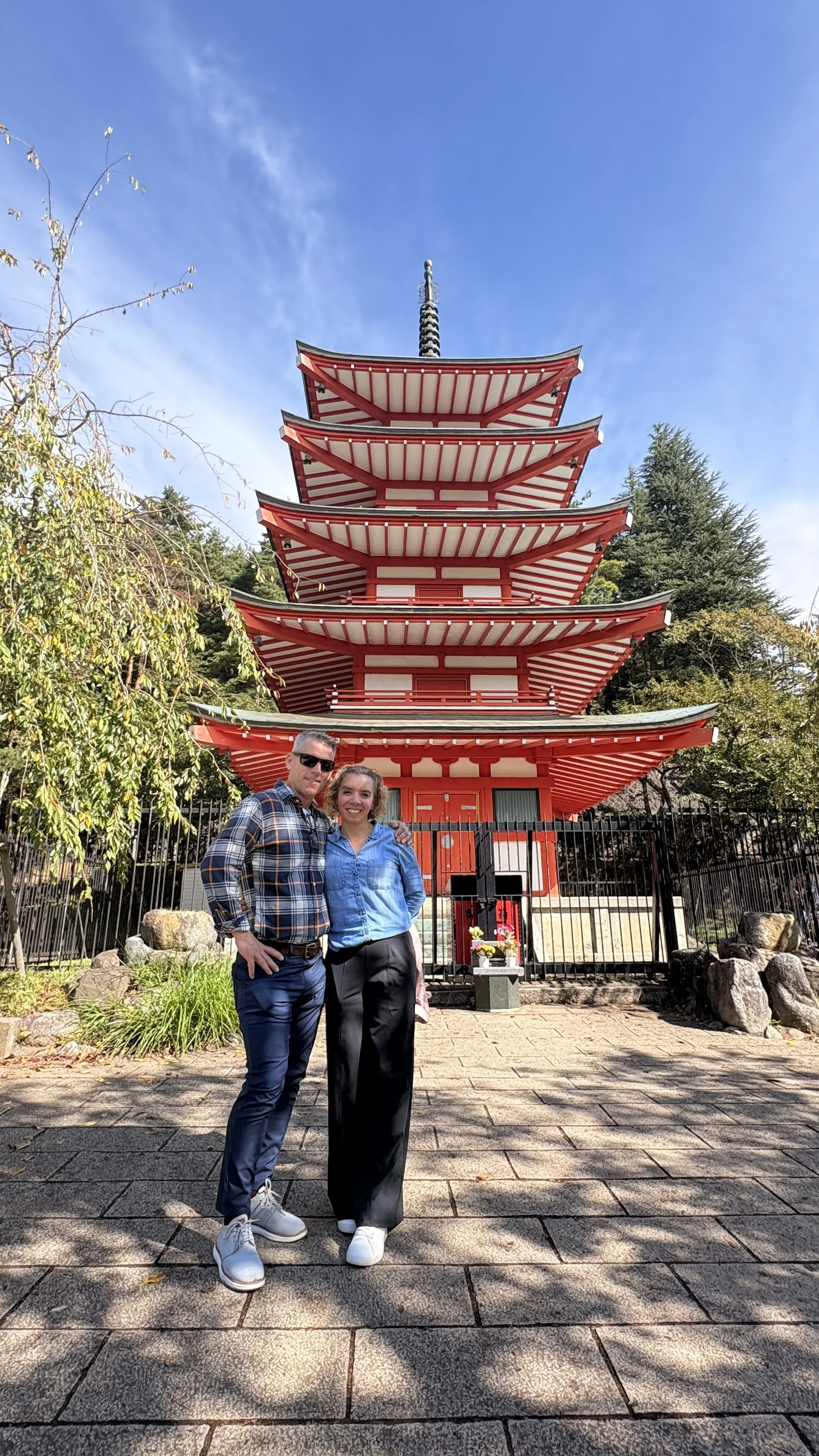 A man and woman standing in front of a traditional Japanese five-story pagoda, smiling and posing for the camera on a sunny day.
