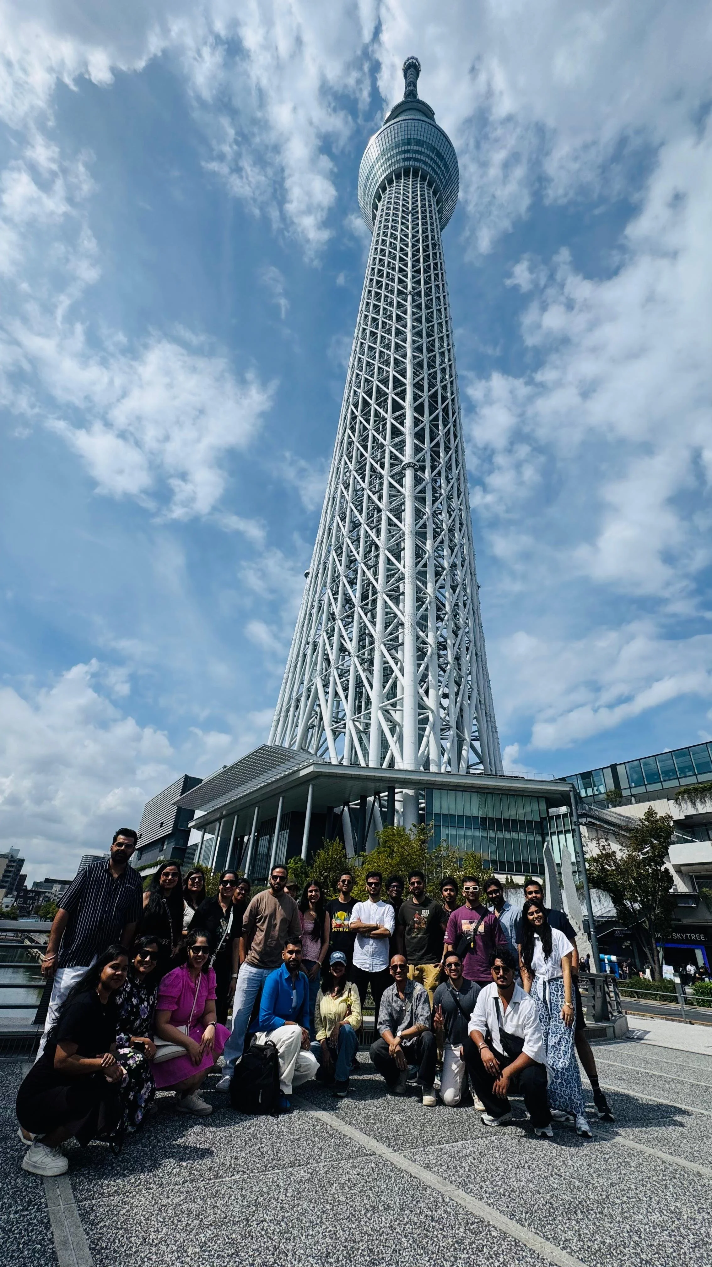 A group of people gathered in front of Tokyo Skytree, a tall, lattice-structured tower with a viewing platform near the top, under a partly cloudy sky.