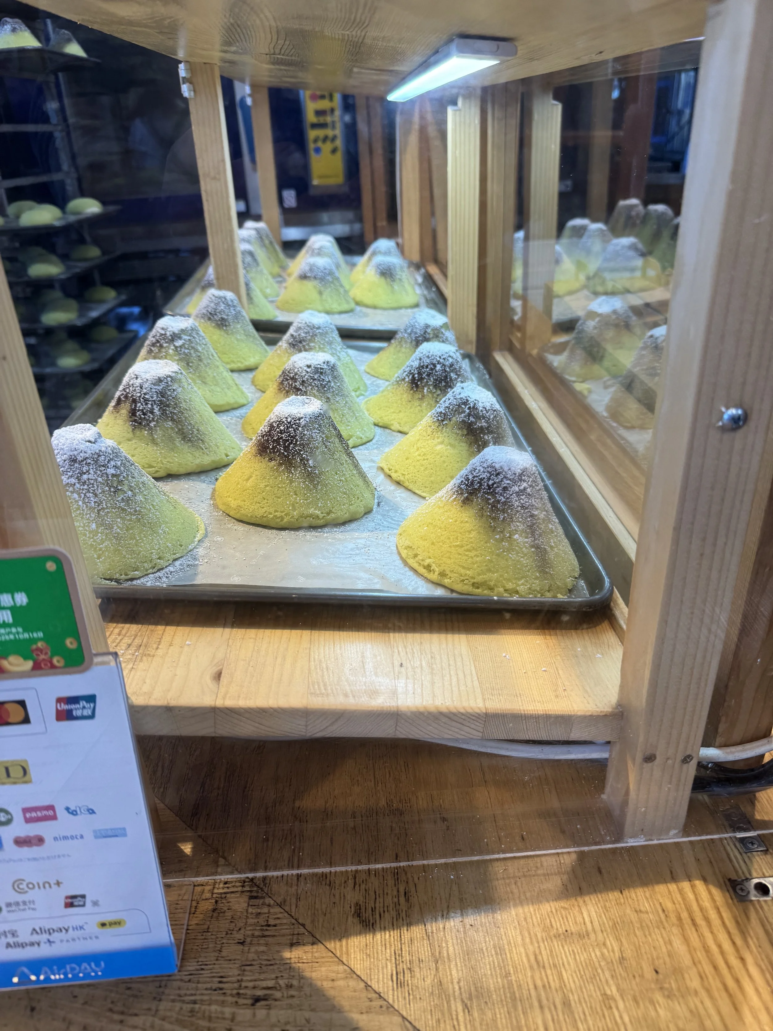 A display case containing several green and brown volcano-shaped cakes dusted with powdered sugar, inside a wooden display with a light source overhead.