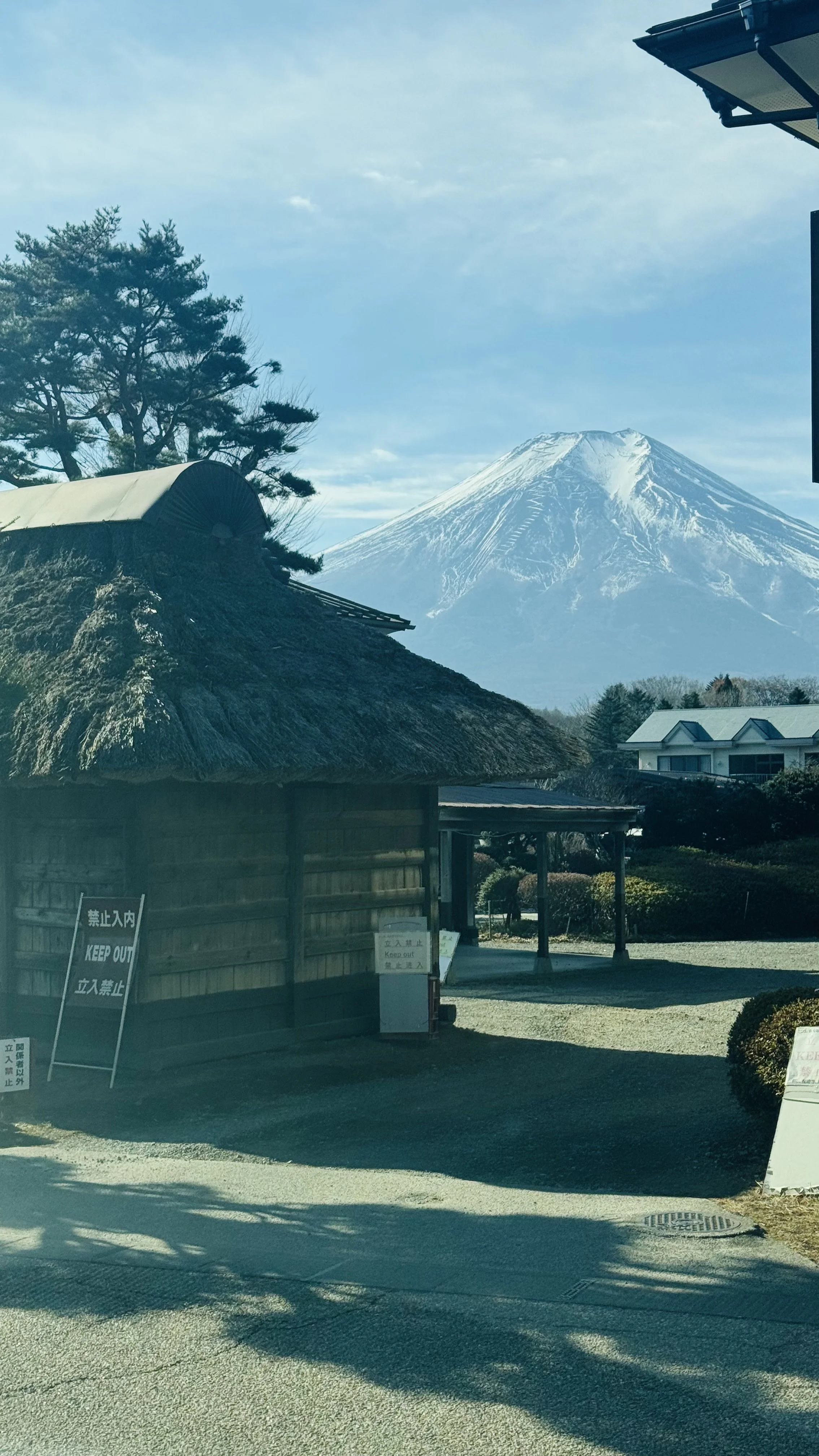 View of Mount Fuji with a traditional thatched-roof building and trees in the foreground, under a partly cloudy sky.