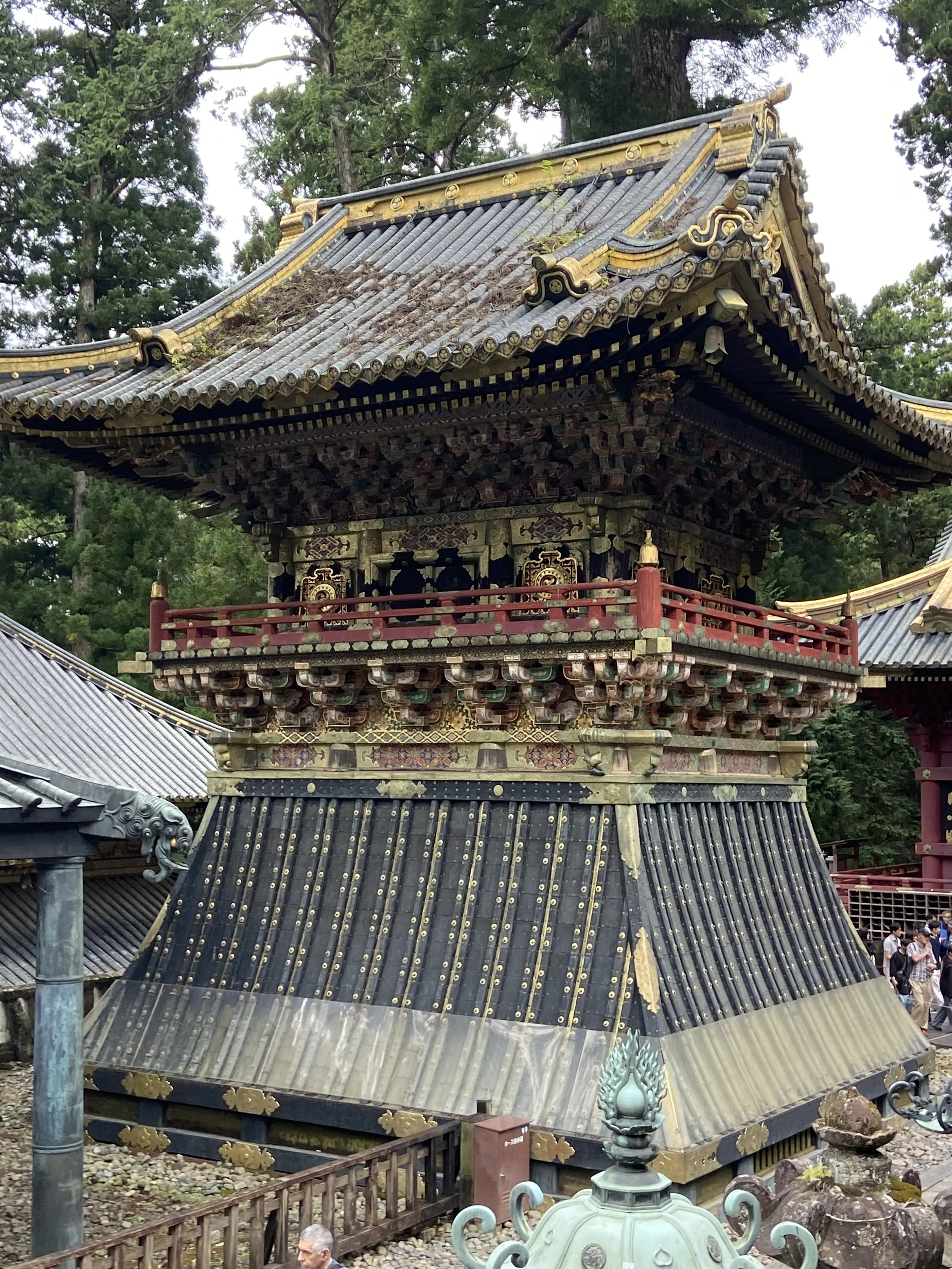 Traditional Japanese temple pagoda with ornate wooden architecture, gold accents, and a guarded staircase surrounded by trees and visitors.
