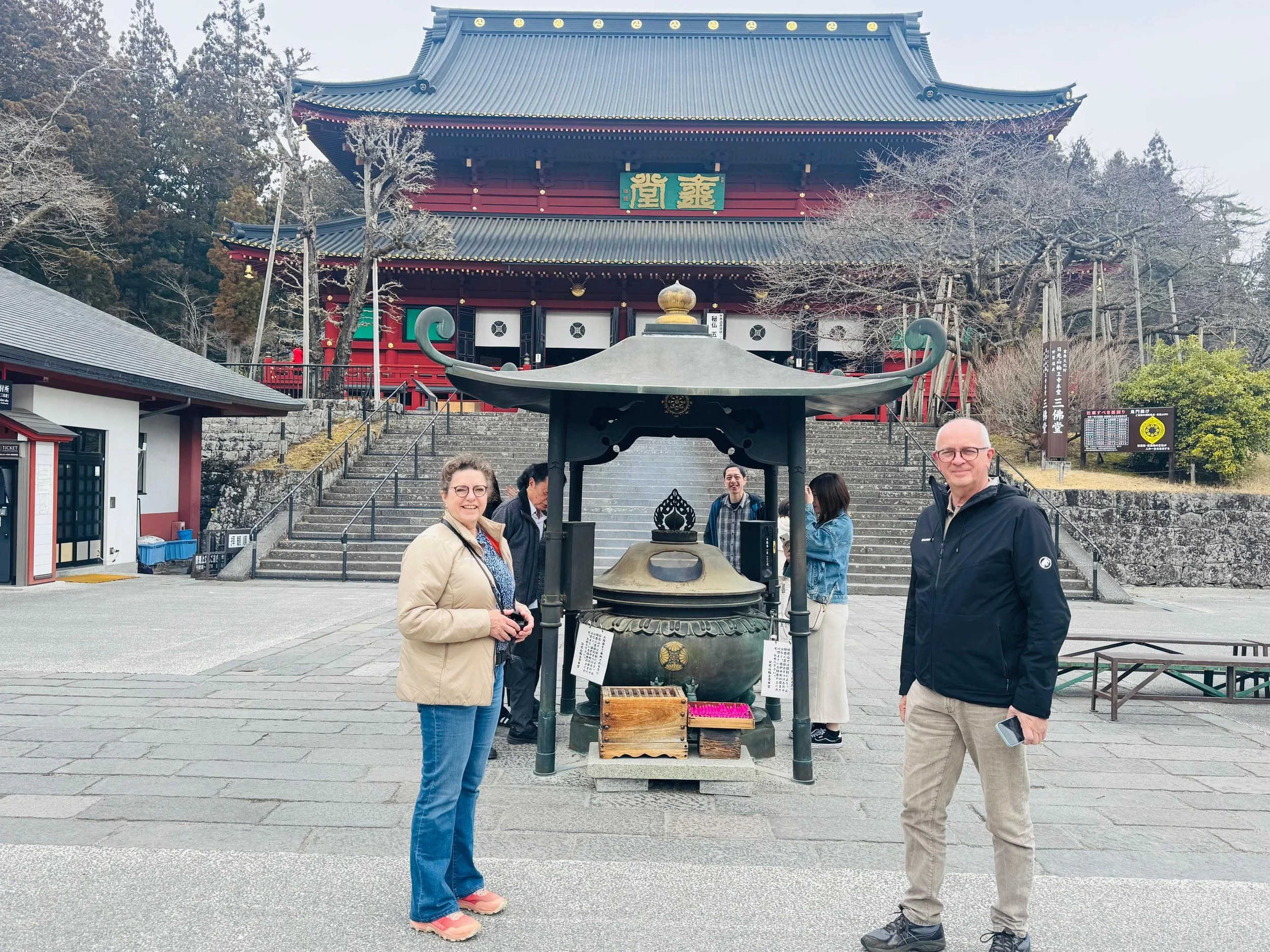 Two people standing in front of a large traditional Japanese temple, with a large bronze incense burner in the foreground, and leafless trees and stairs behind them.