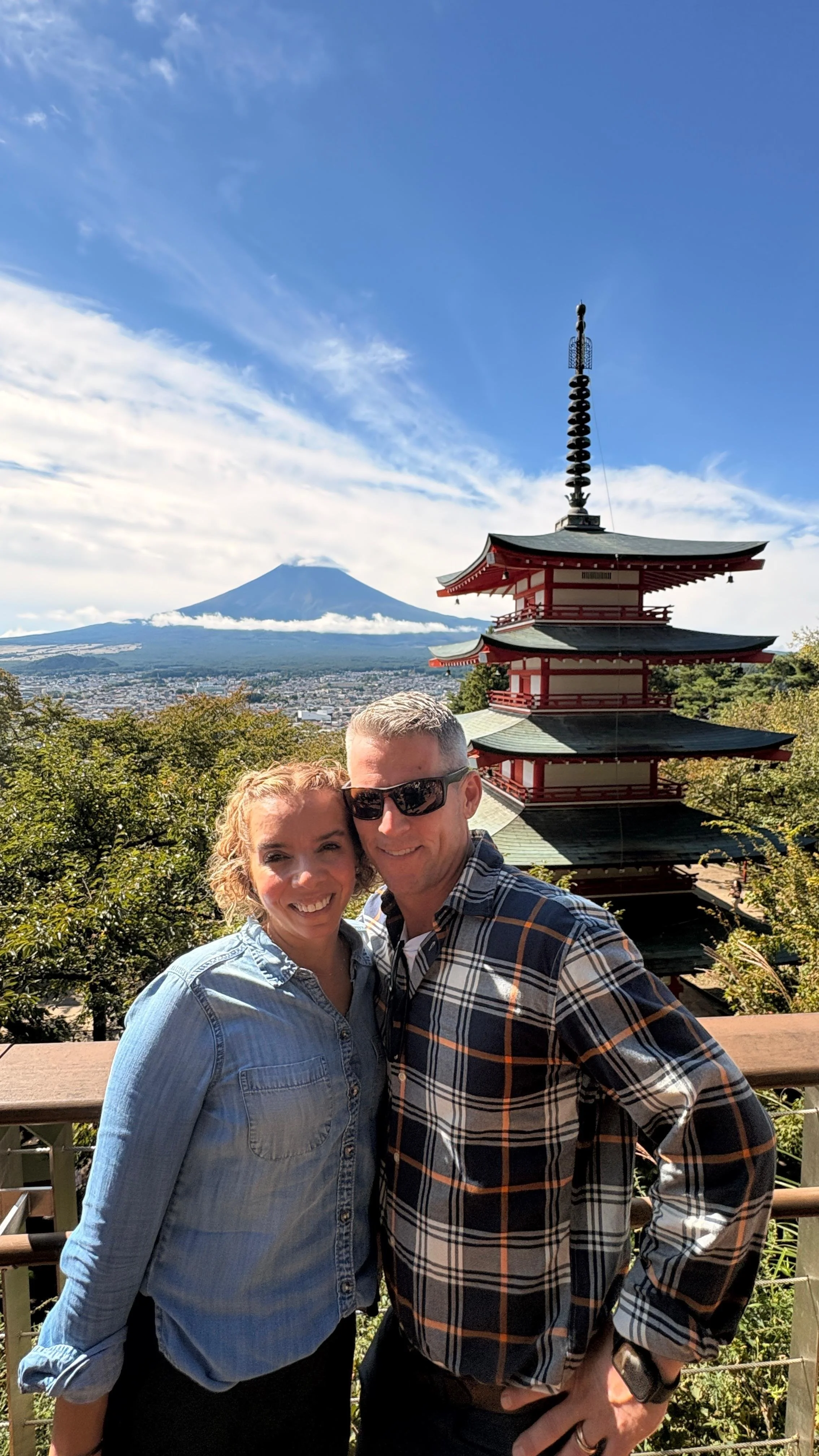 A smiling couple taking a selfie with Mount Fuji in the background, a traditional Japanese pagoda, and lush greenery under a clear blue sky.