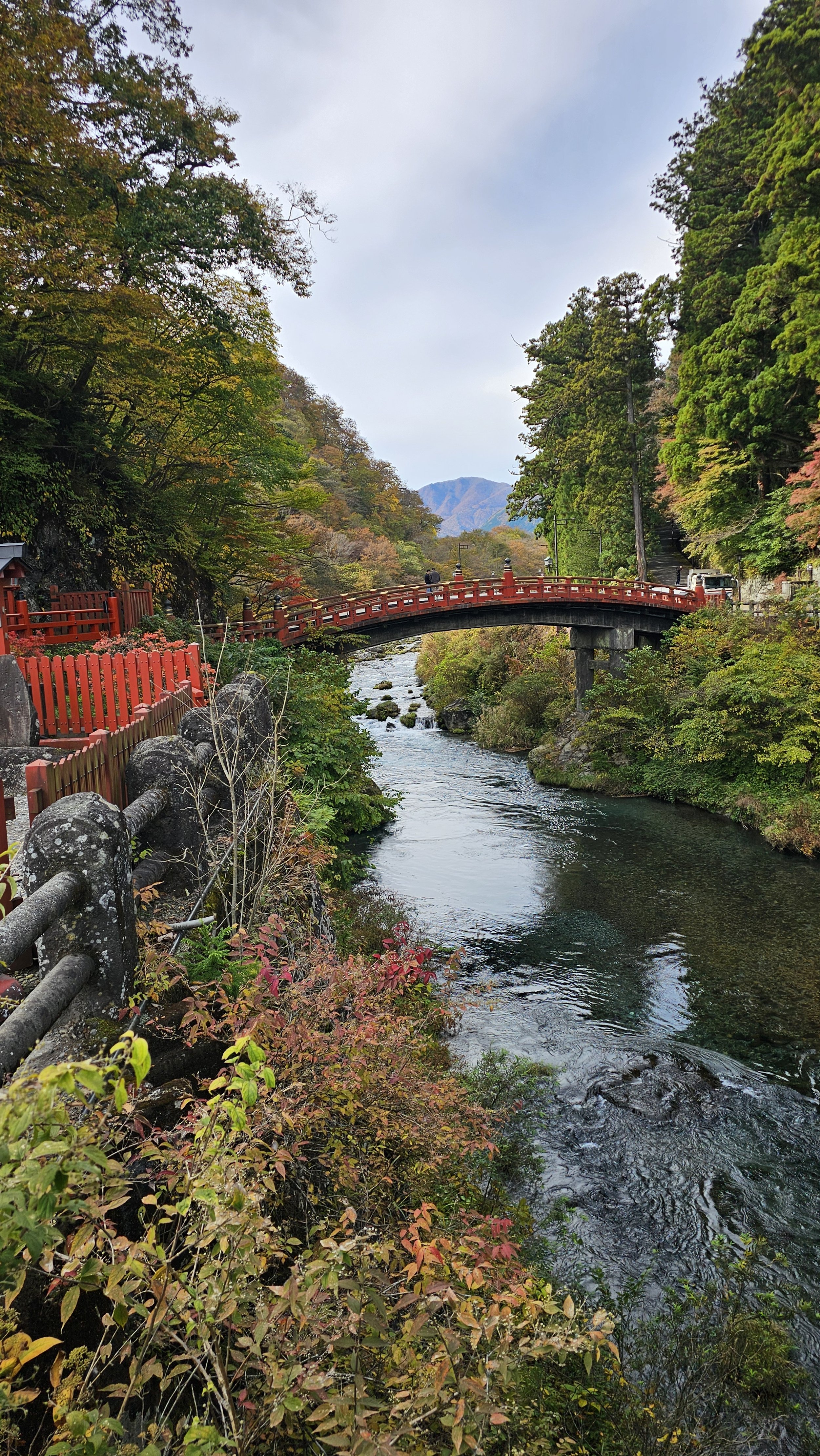 A scenic river view with a red bridge arches over the water, surrounded by lush greenery and trees with some fall foliage, and distant mountains in the background.