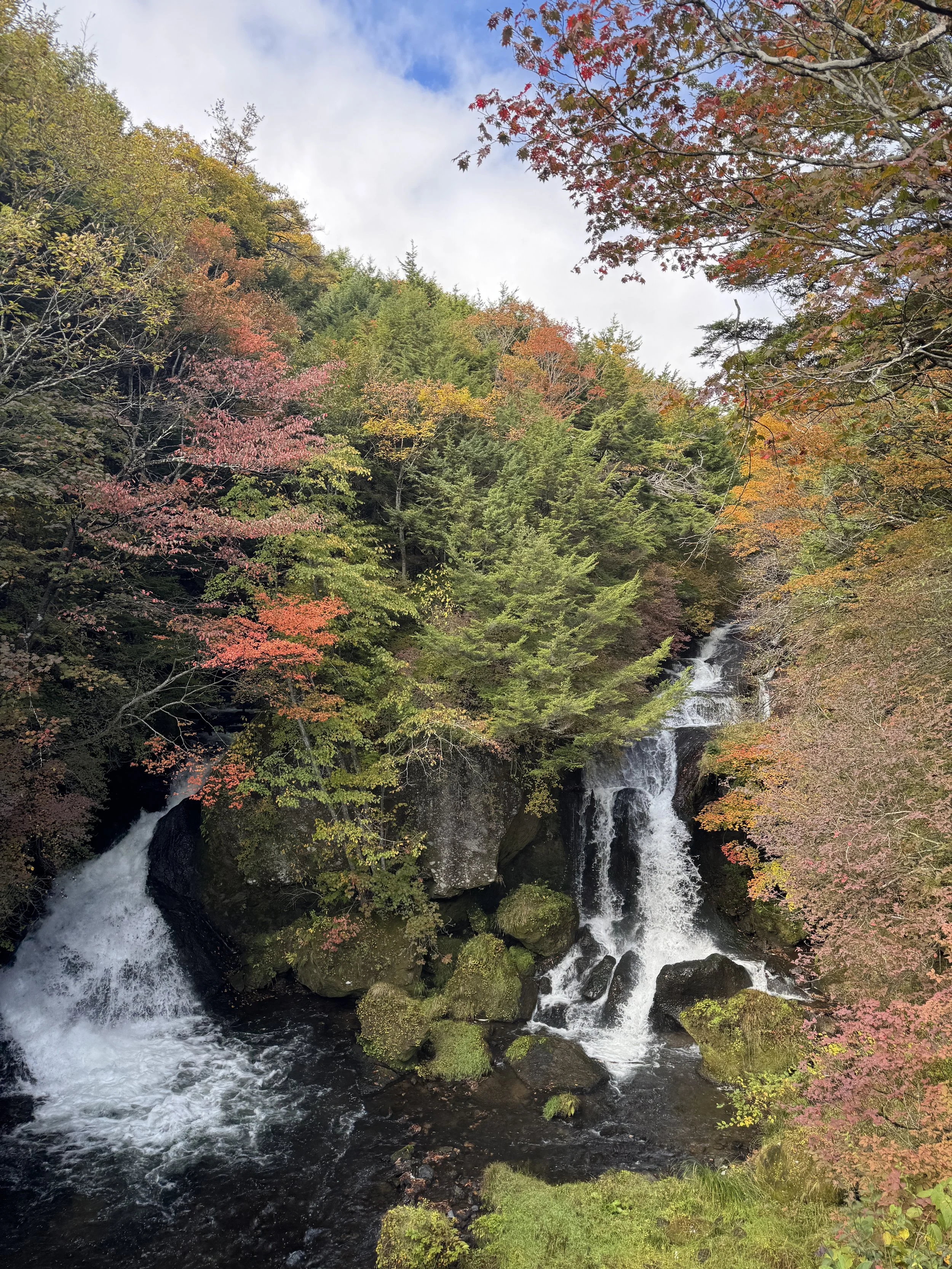 Scenic waterfall surrounded by colorful autumn trees with red, orange, yellow, and green leaves in a lush forest.