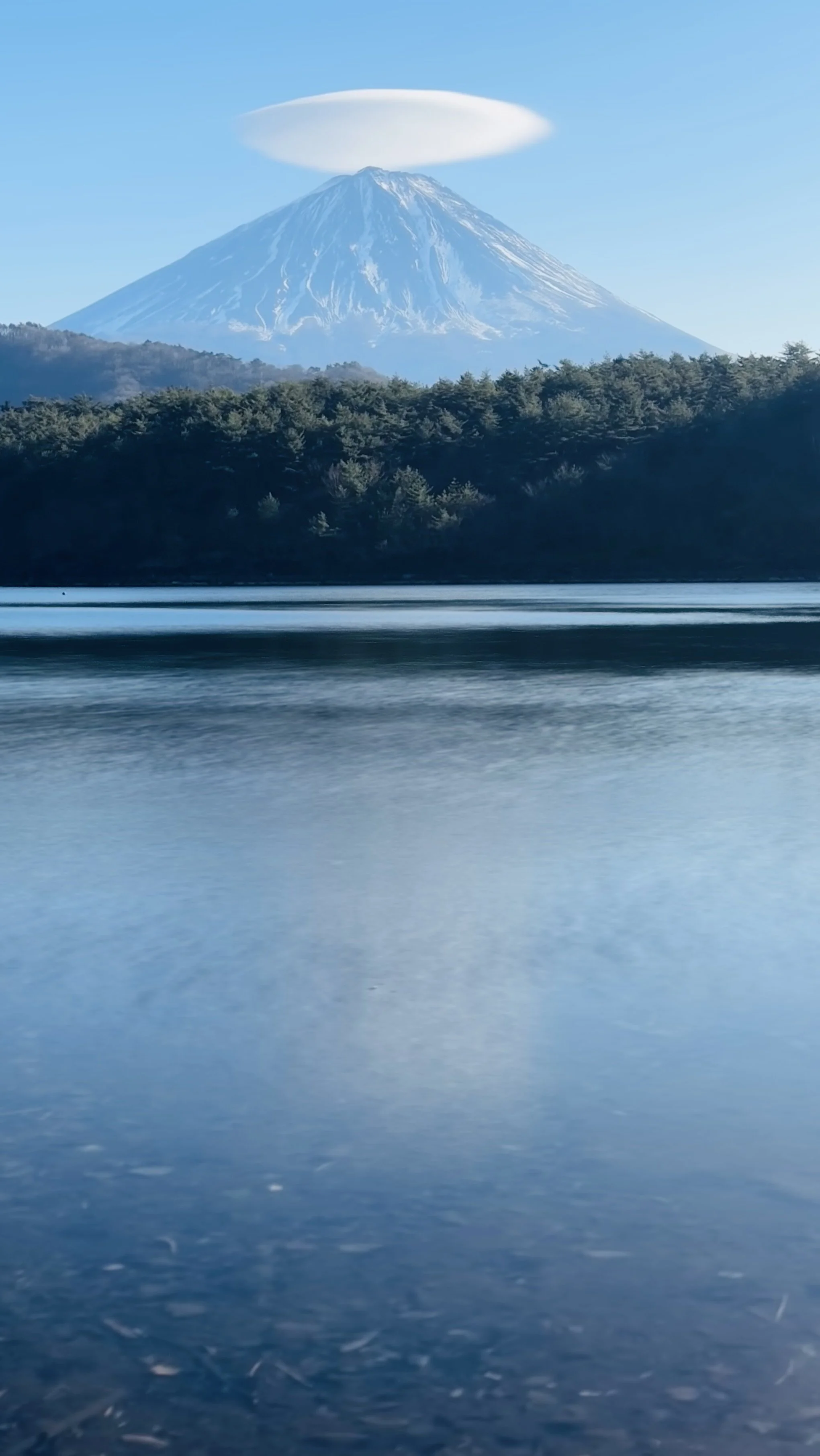 Snow-capped mountain with a halo-shaped cloud above its peak, seen across a body of water with forested shoreline.