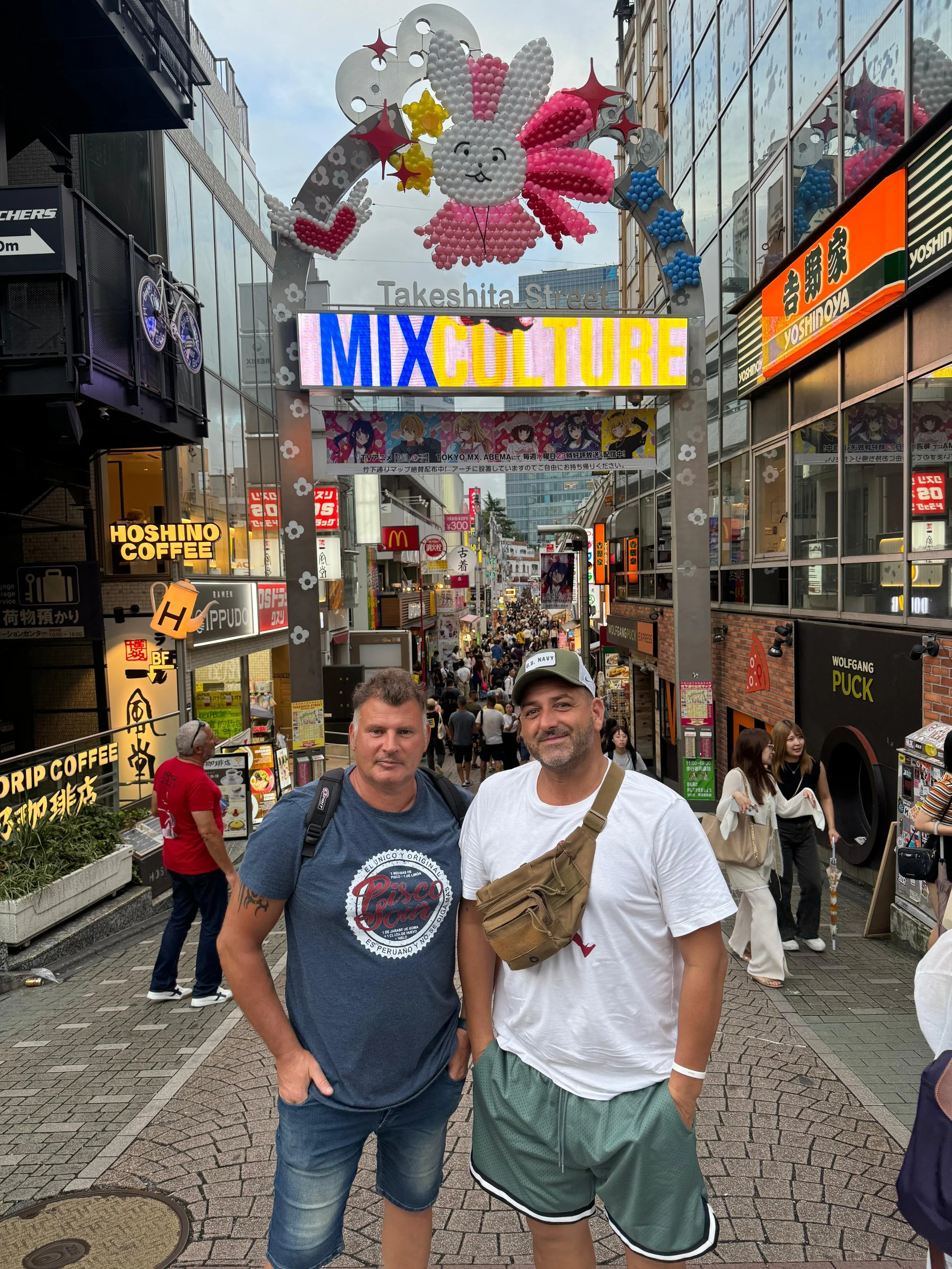 Two men standing on Takeshita Street in Harajuku, Tokyo, Japan, posing for a photo. The street is busy with pedestrians and has many colorful shop signs, including a large inflatable bunny decoration hanging above with the sign 'MIX CULTURE'.